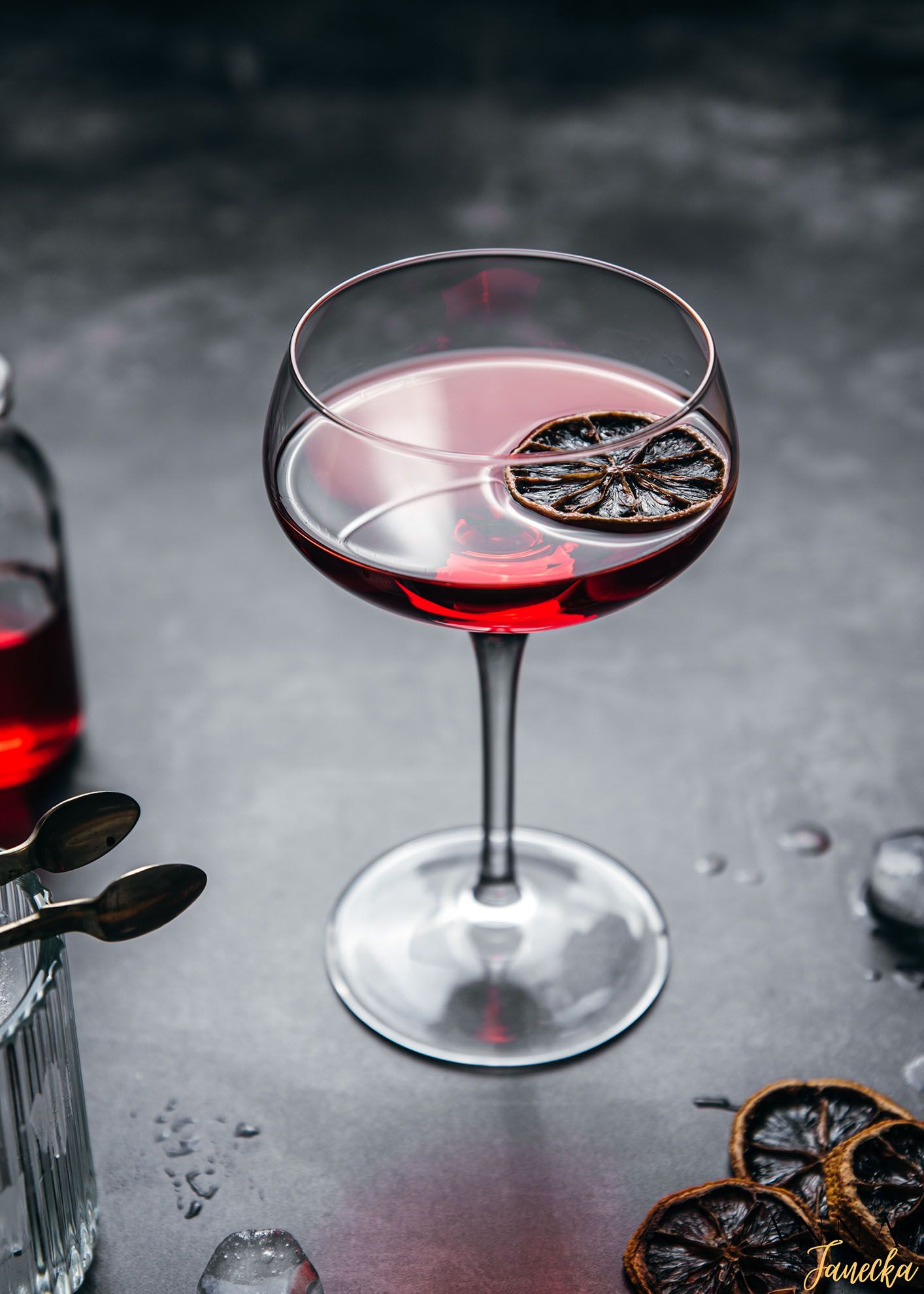 Red cocktail in a coupe glass garnished with a dried citrus wheel on dark background.