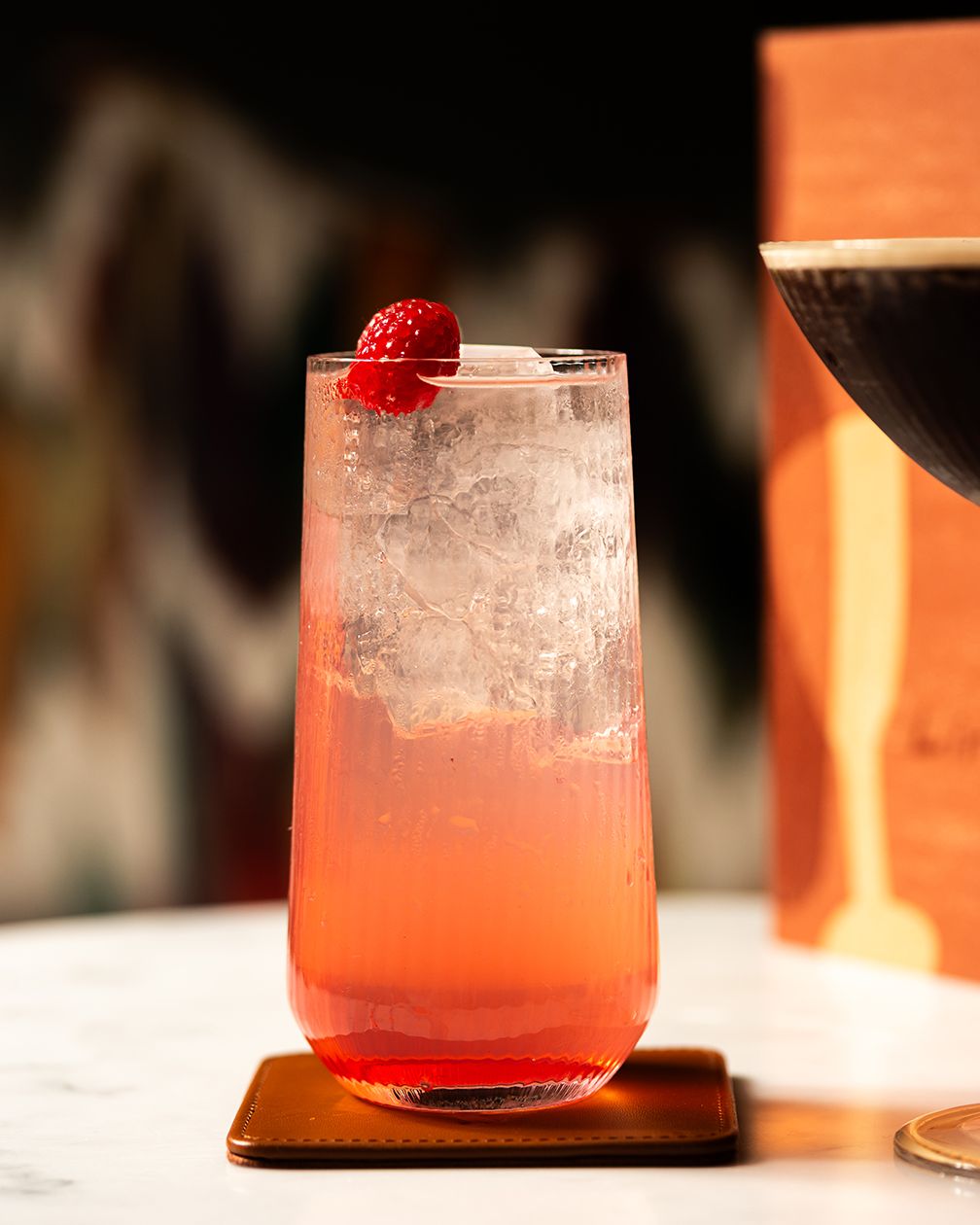 A vibrant pink raspberry cocktail in a tall glass with crushed ice, garnished with fresh raspberries on a wooden coaster.