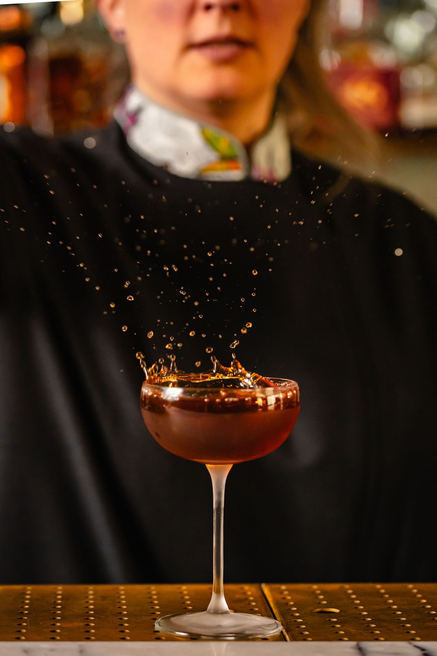 Bartender presents a dark cocktail in a coupe glass with dramatic liquid splash against a warm bar backdrop.