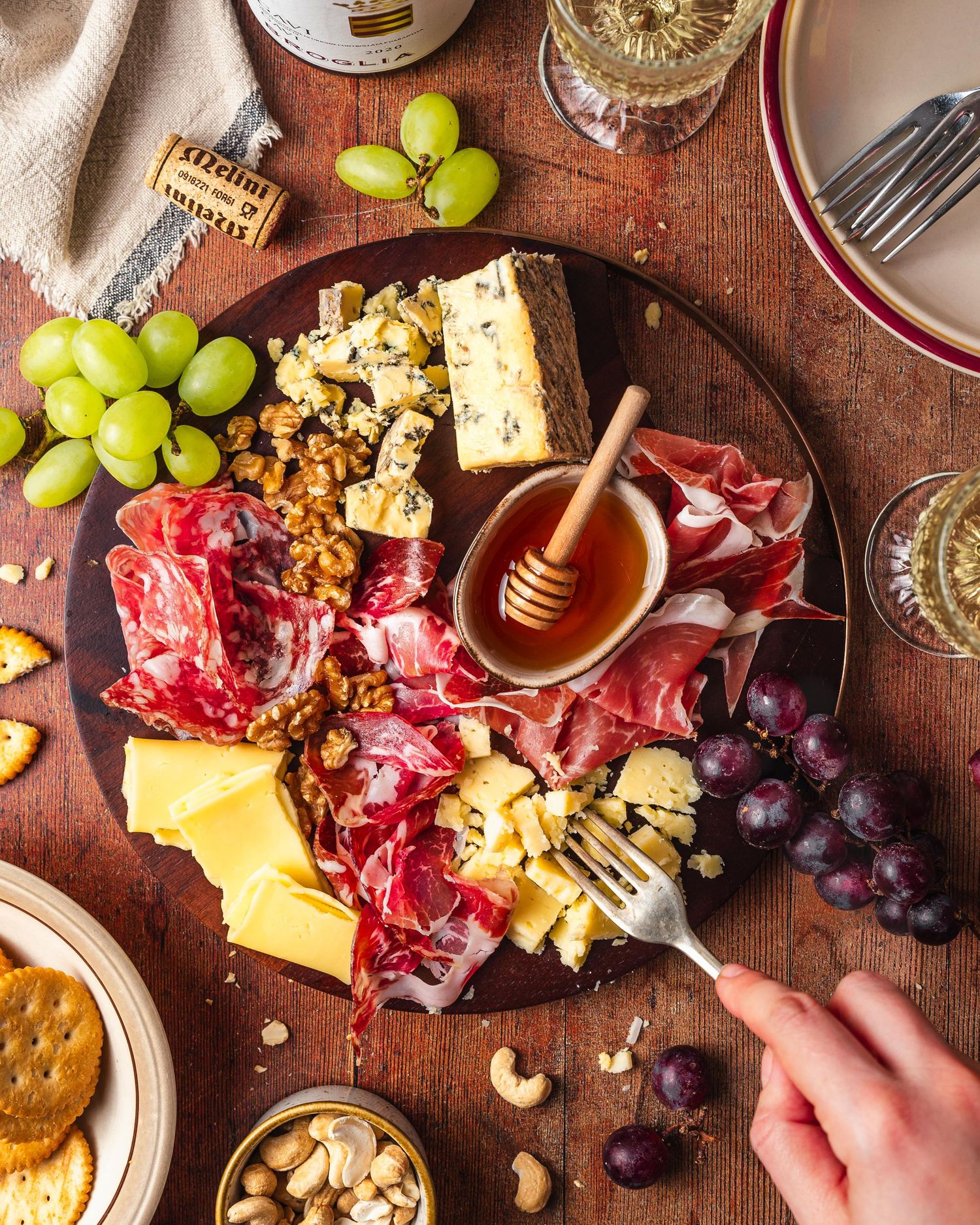 A charcuterie board with cured meats, cheeses, grapes, walnuts, honey, and crackers on a wooden table.