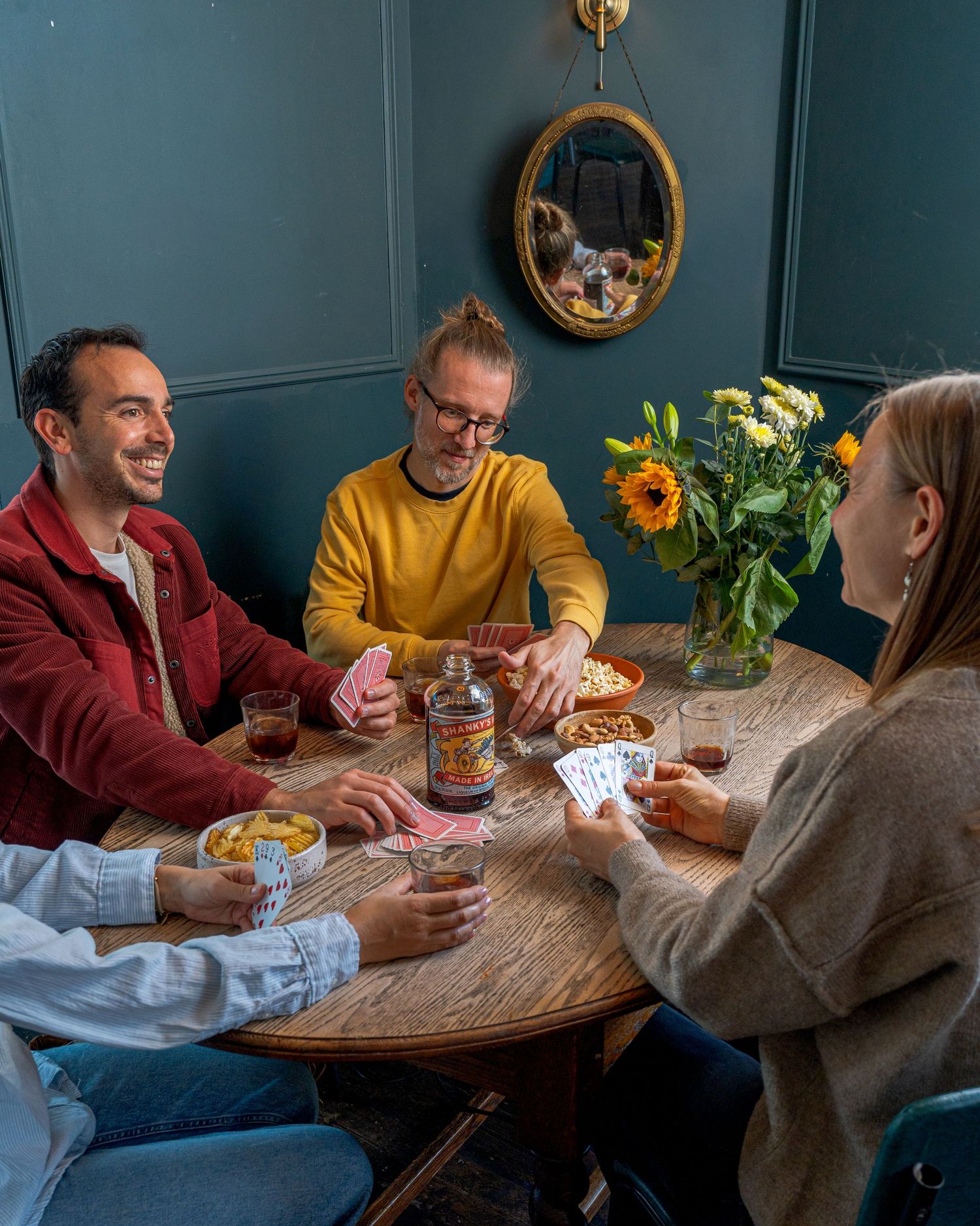 A group playing cards around a wooden table in a room with teal walls and sunflowers in the background.