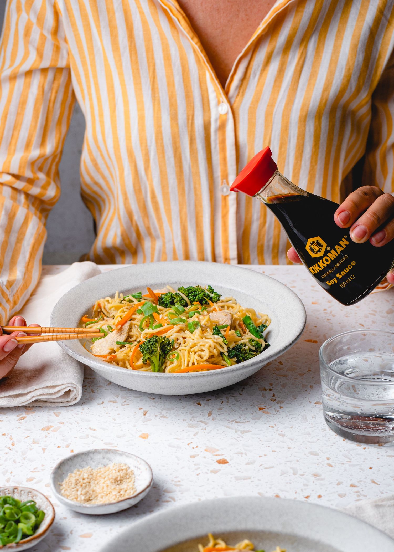 Bowl of pasta being served with garnishes and condiments on white table surface.