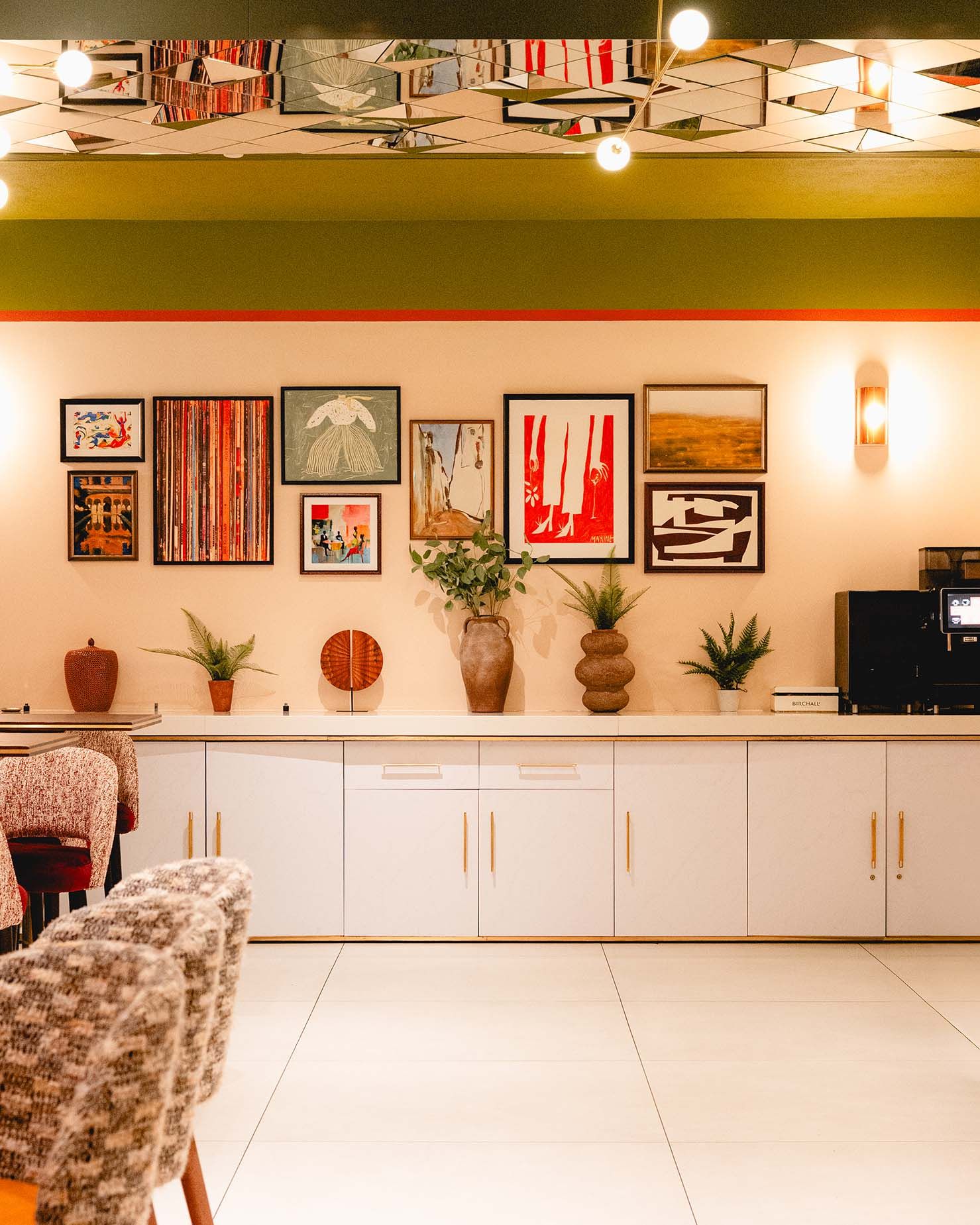 Eclectic hotel lobby with gallery wall art, white credenza, potted plants, and retro decor under green ceiling.