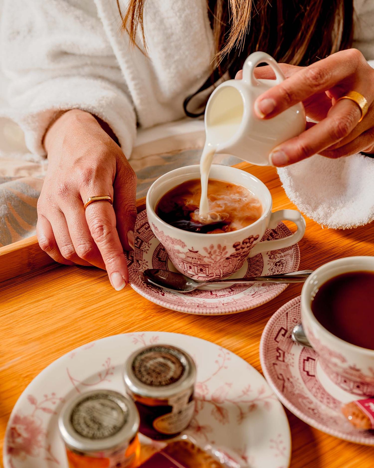 Woman in white robe pouring cream into floral teacup on wooden tray with spoons and small jars.