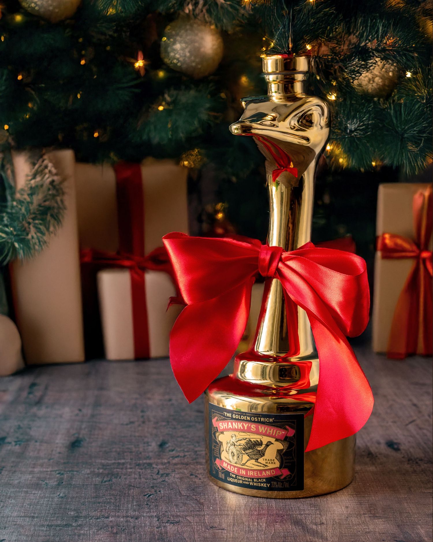 Gold ostrich-shaped liquor bottle with red bow displayed in front of Christmas tree with gifts.