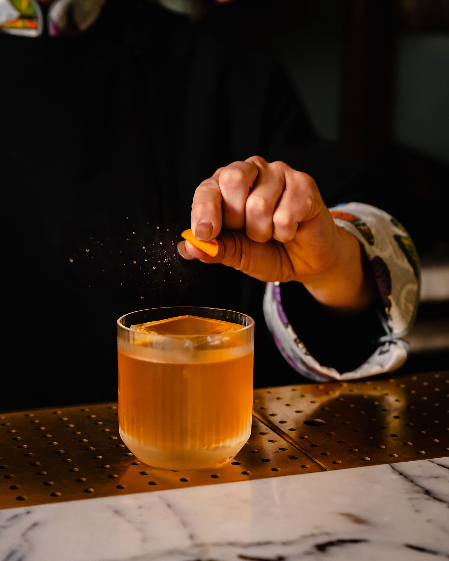 Bartender squeezing fresh orange peel over an amber cocktail in a rocks glass on a marble bar.
