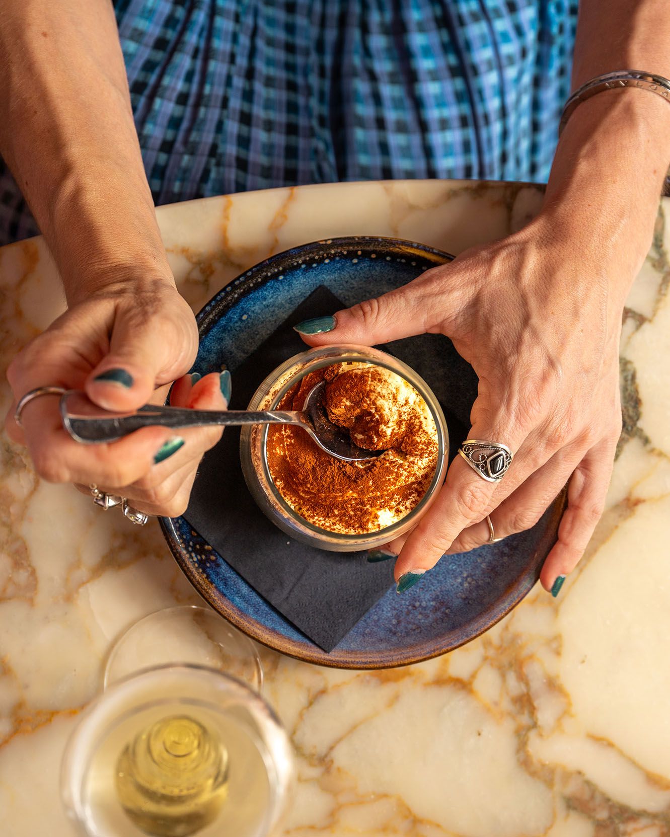 Hands scooping a spiced dessert from a small jar on a blue ceramic plate, with a glass of white wine nearby.