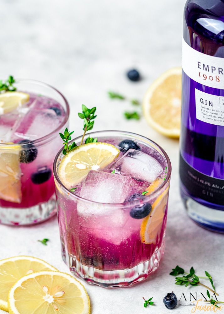 Purple gin cocktails garnished with lemon slices, blueberries and fresh herbs in crystal glasses.