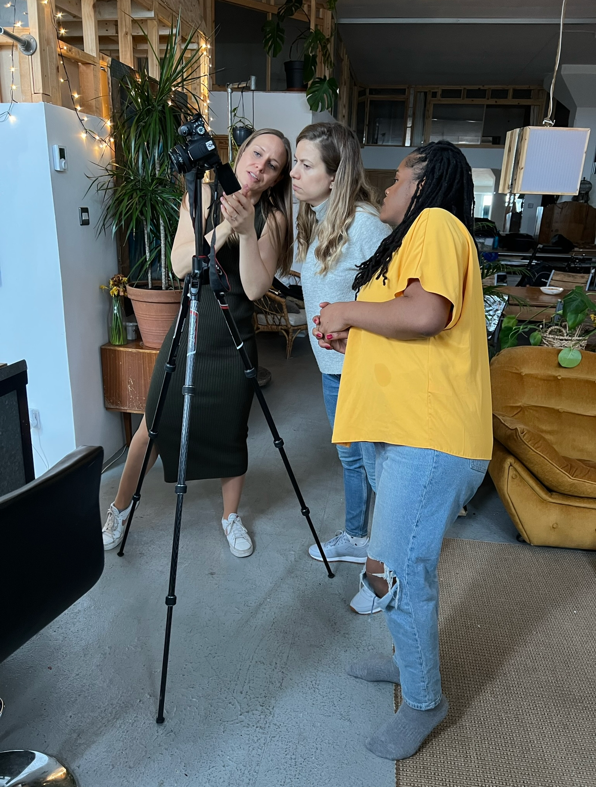 Three people gather around a camera on a tripod in a room with houseplants and casual decor.