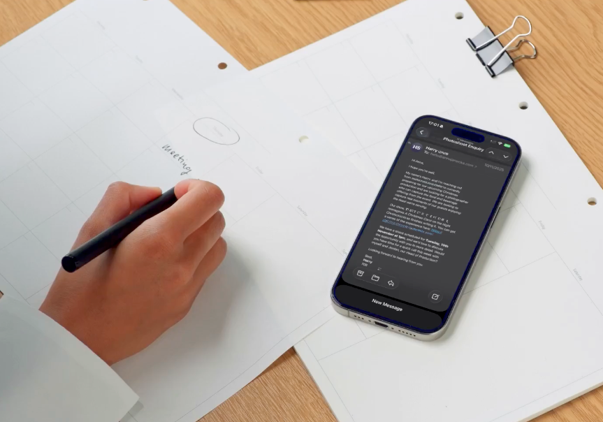 A hand writing on white paper next to a smartphone displaying text on a wooden desk.