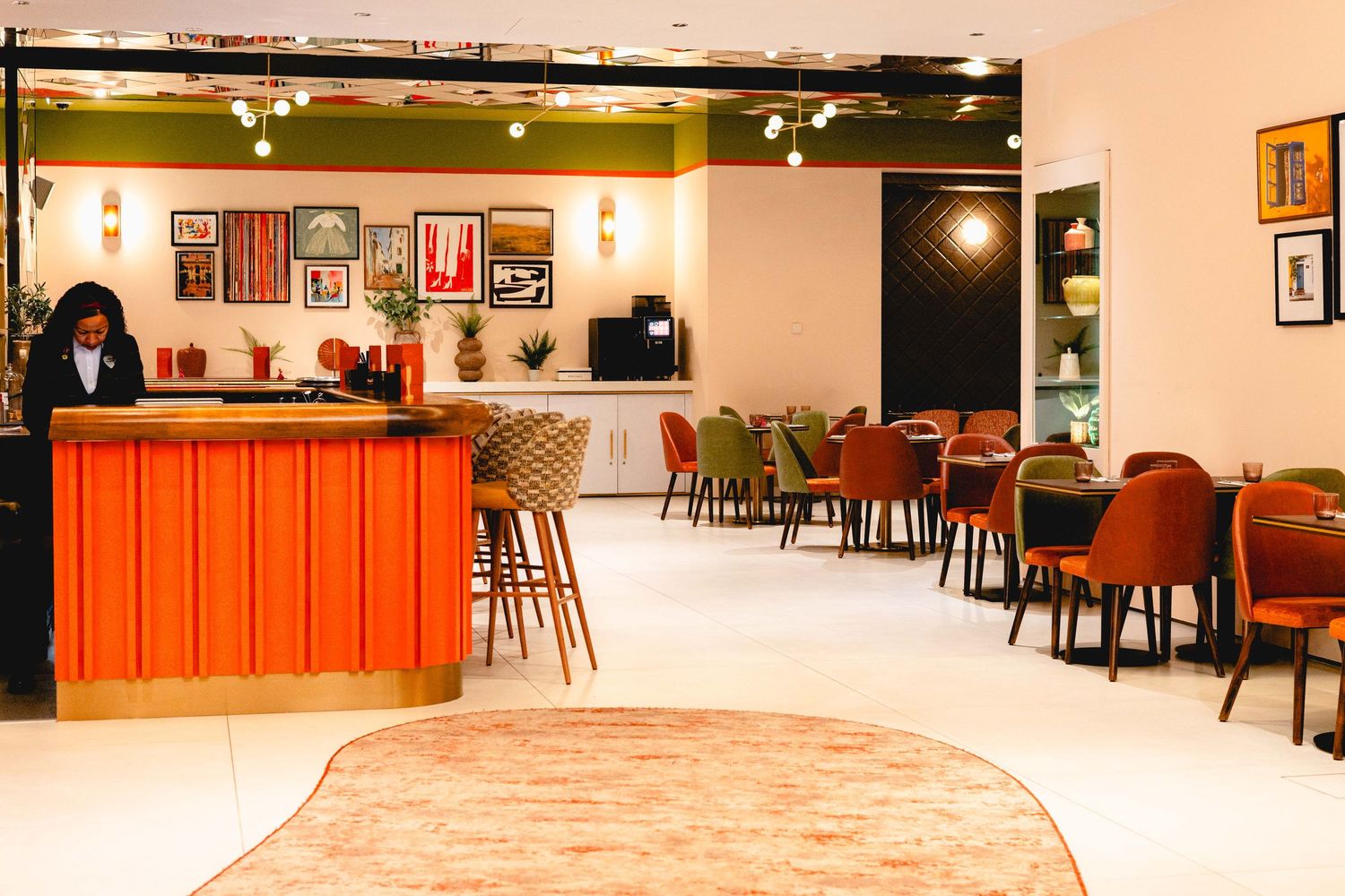 Modern hotel lobby with orange reception desk, colorful artwork, and eclectic mix of green and rust dining chairs.