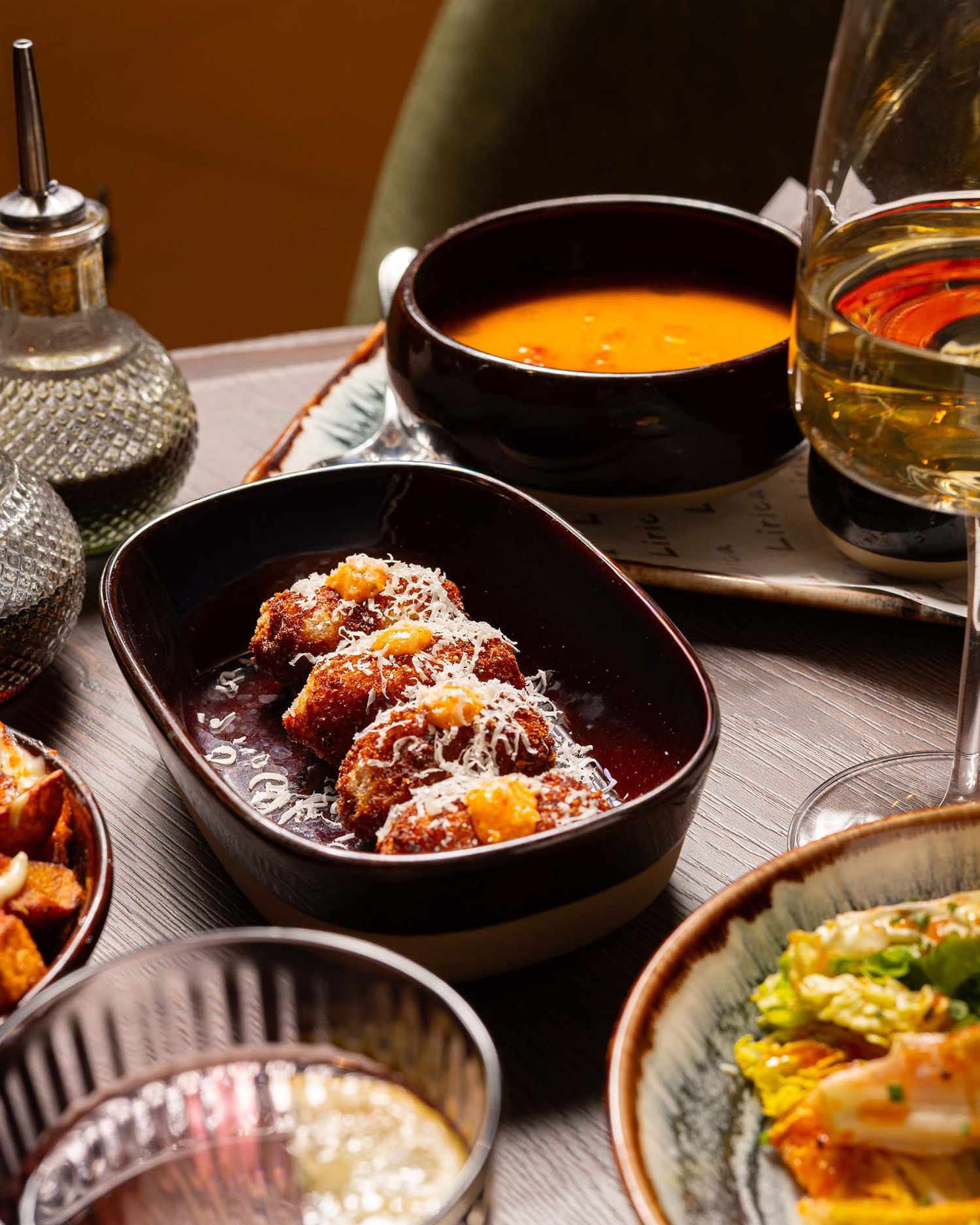 A table spread with tapas-style dishes, including meatballs in sauce, tomato soup, and garnished plates with wine glasses.