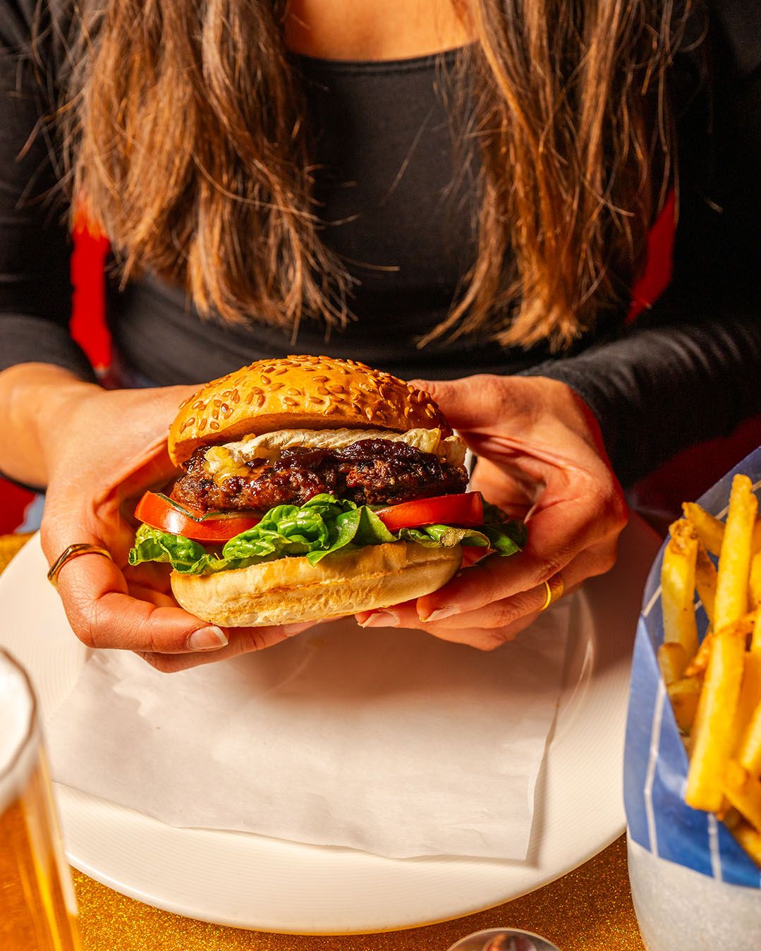 Woman holding a juicy burger with lettuce, tomato, and beef patty on sesame bun, served with fries and beer.