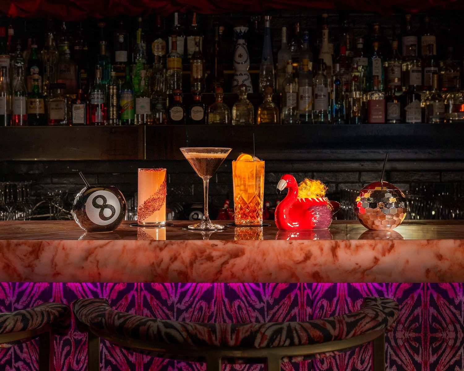 Four colorful cocktails lined up on a vibrant pink bar counter with a fully stocked backlit liquor shelf behind.