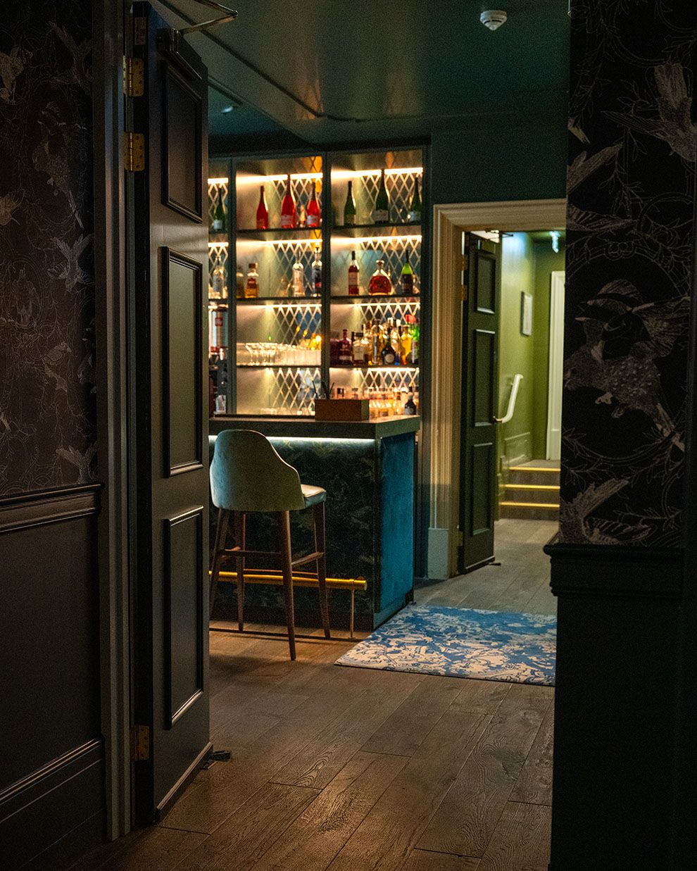 Elegant dimly lit bar with backlit shelves of colorful bottles, teal velvet stools, and dark moody interior design.