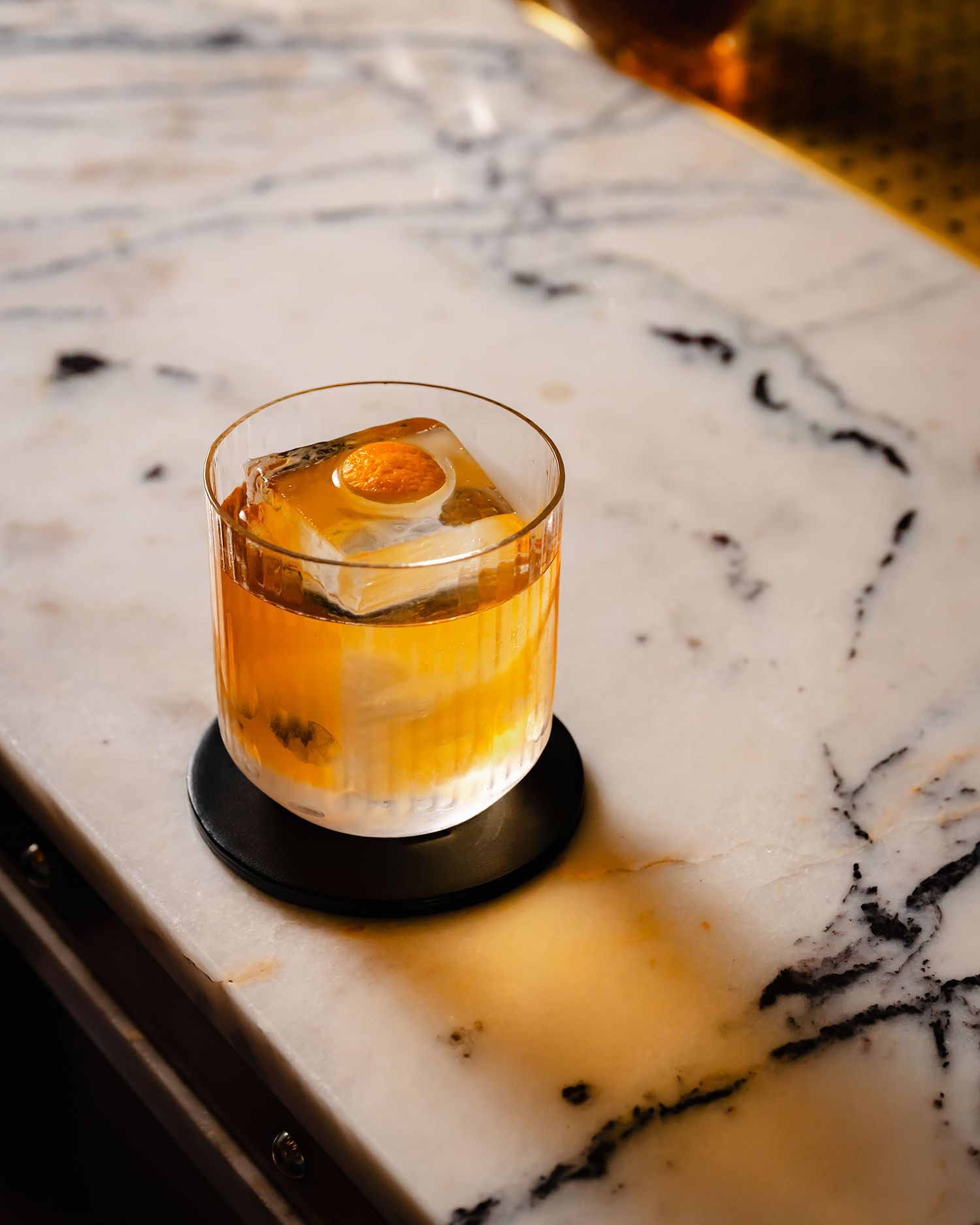 Golden whiskey cocktail with ice cube and orange peel garnish on a black coaster on marble bar.