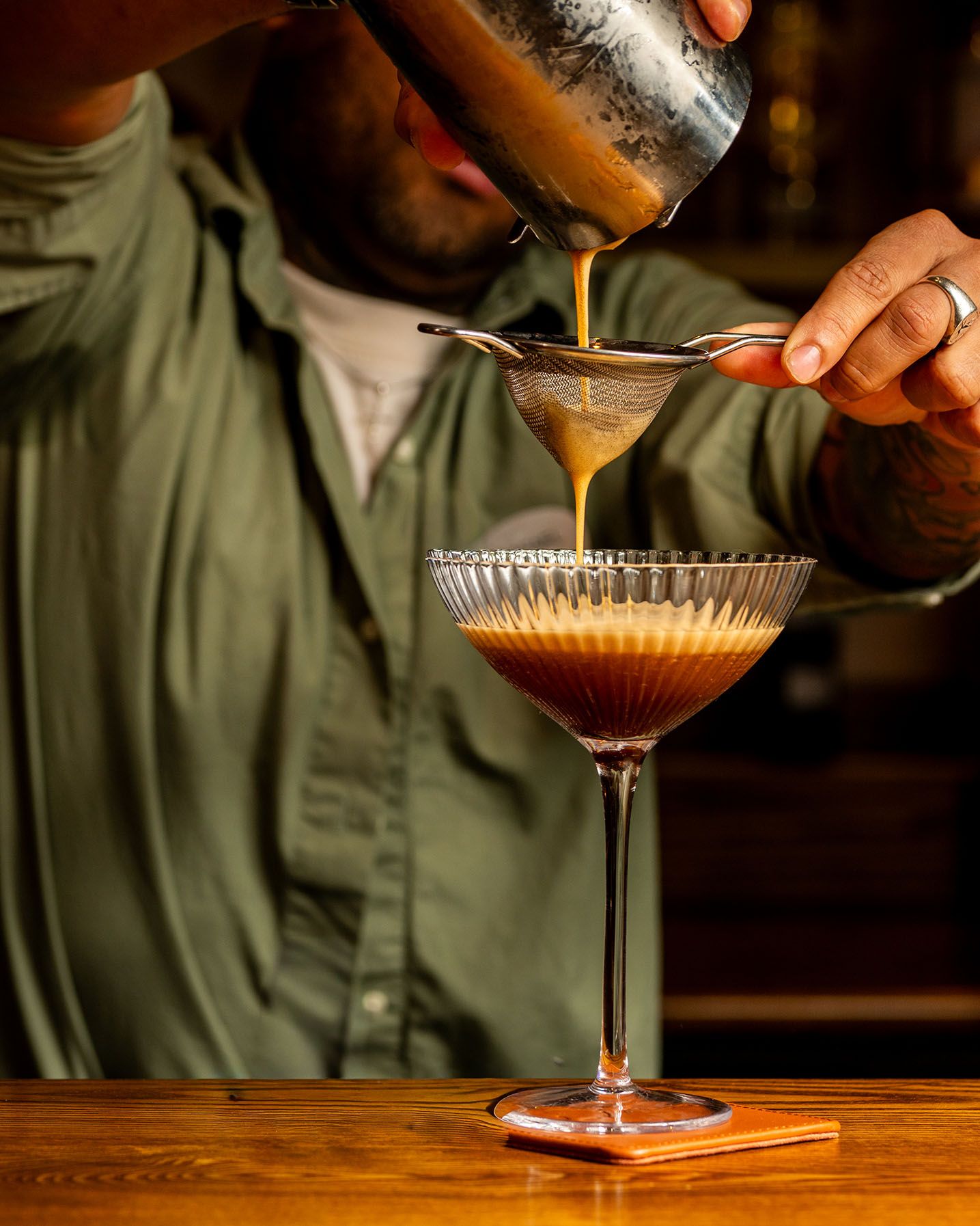 Bartender pouring espresso martini through strainer into elegant coupe glass on wooden bar.