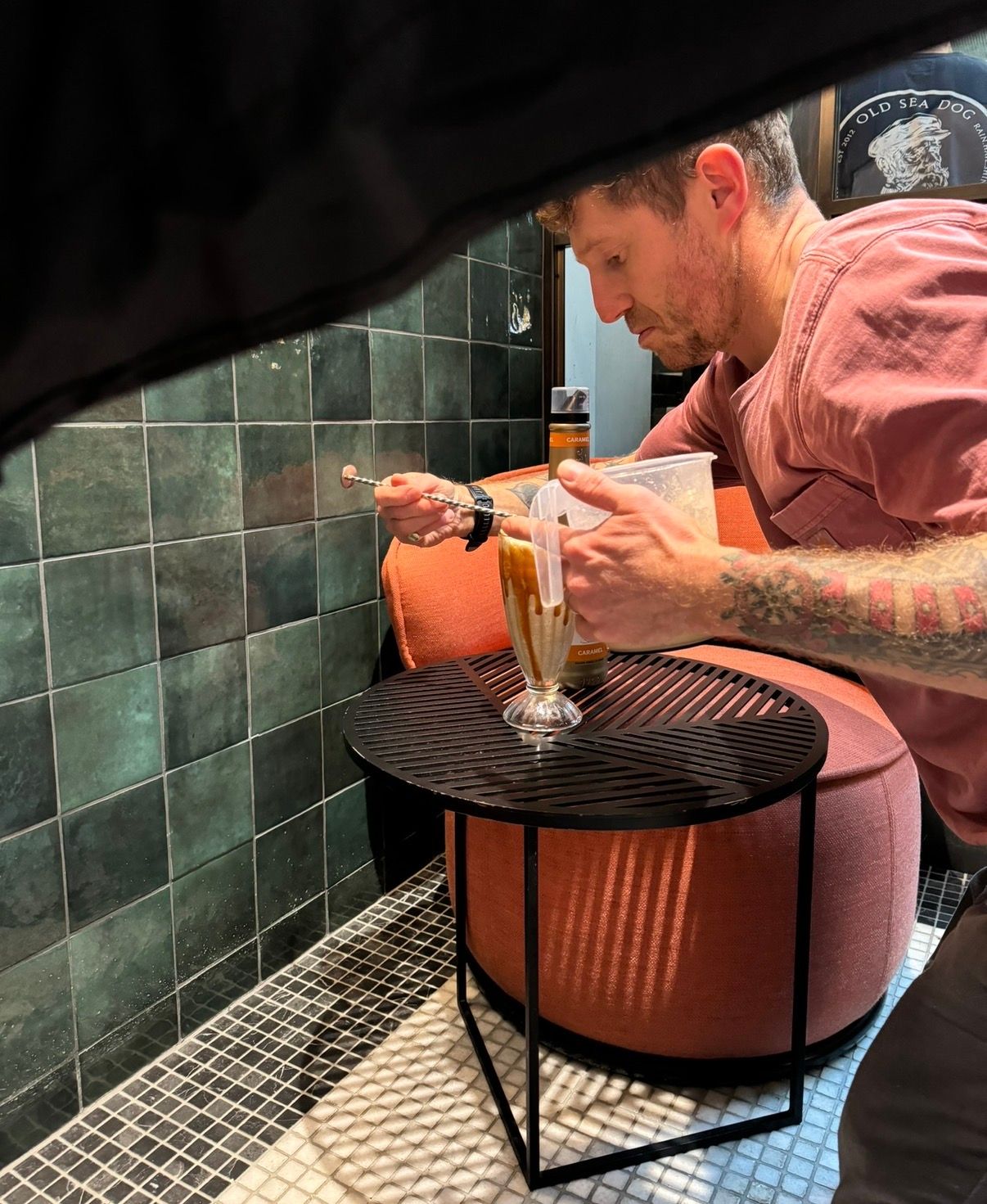 A person in a pink shirt carefully pours ingredients into a glass on a round black table with teal tile walls in background.