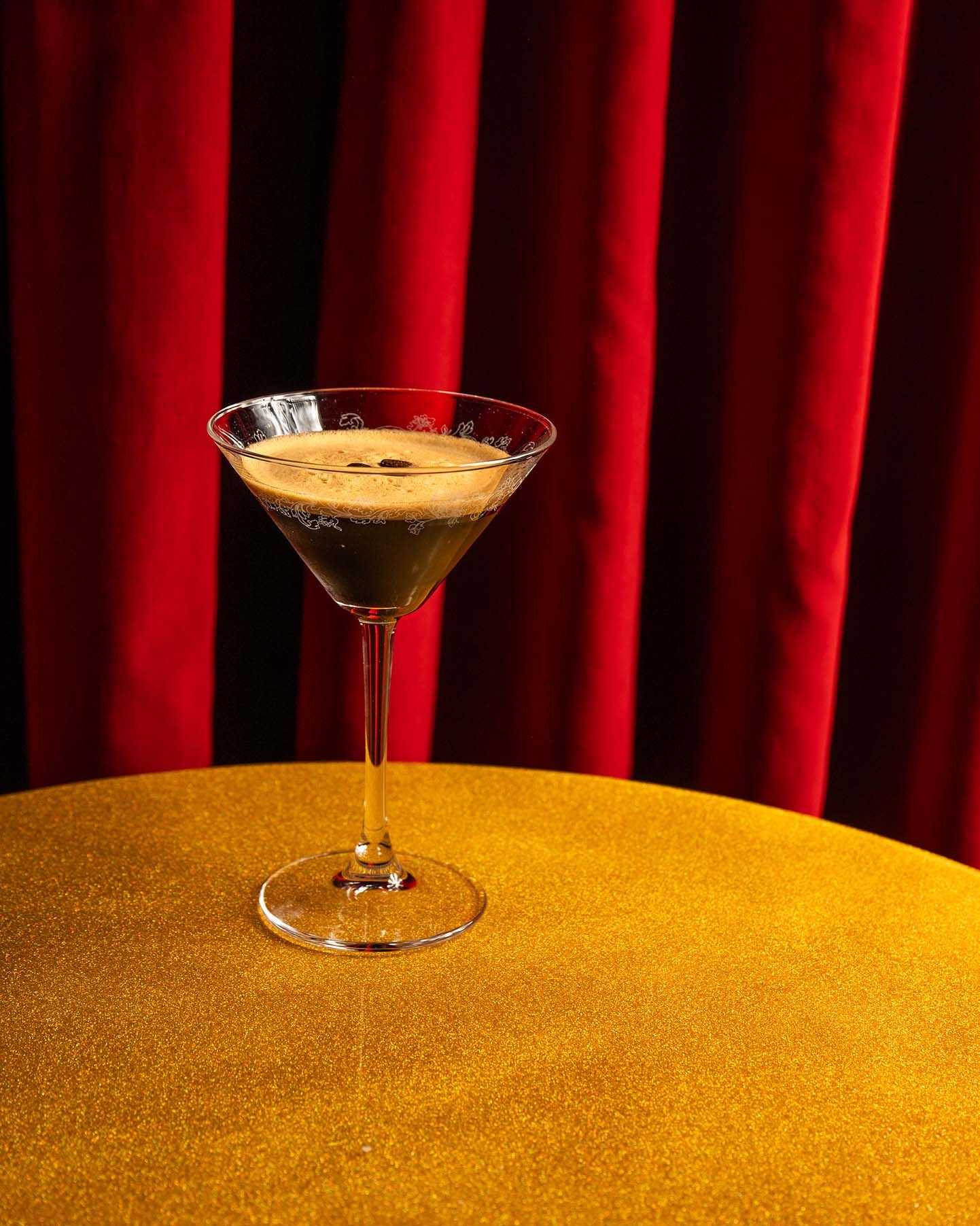 Espresso martini in a classic martini glass on a gold table against a luxurious red velvet curtain backdrop.