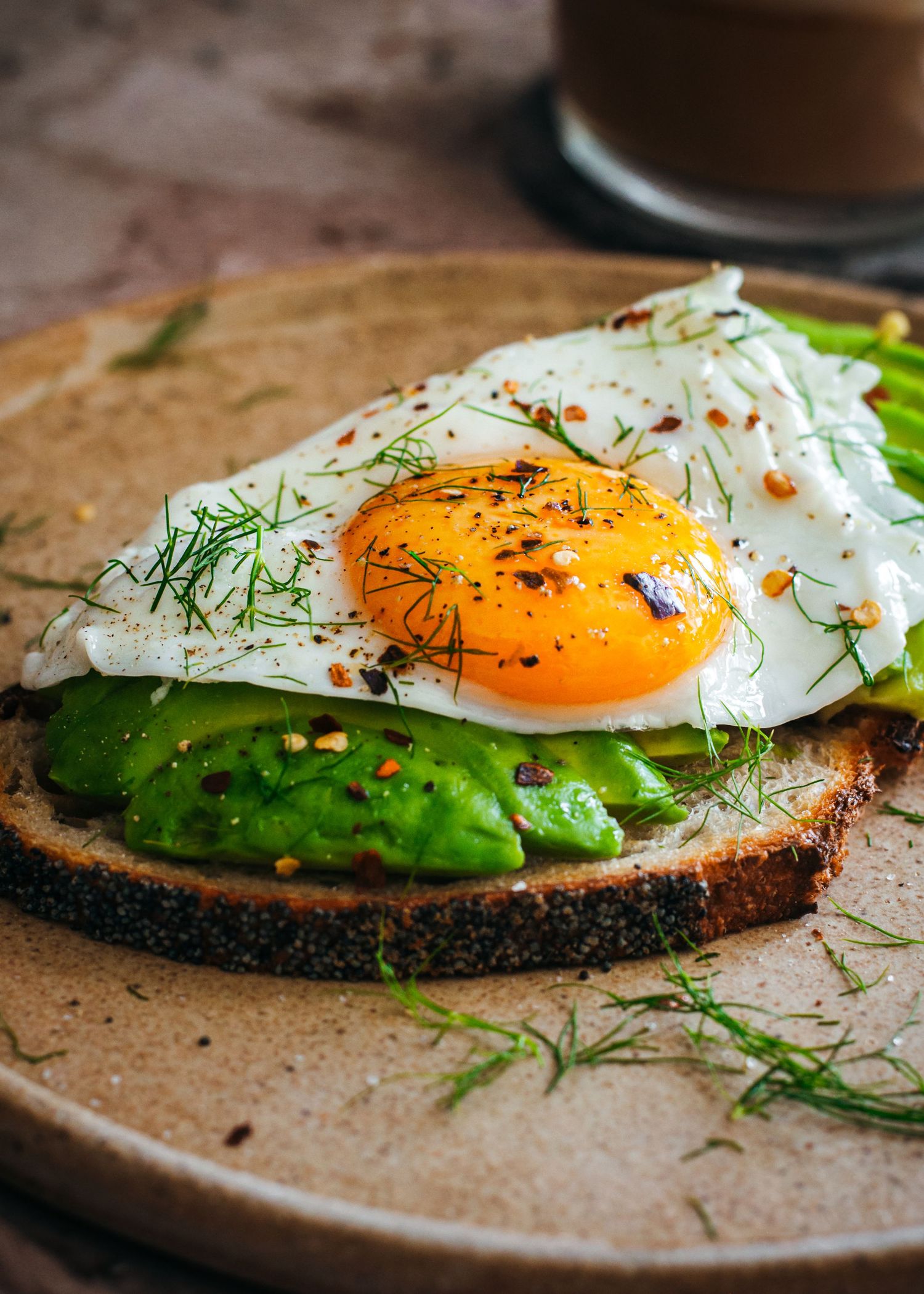 Rye bread topped with mashed avocado, fried egg, and fresh dill garnish served on a rustic brown plate.