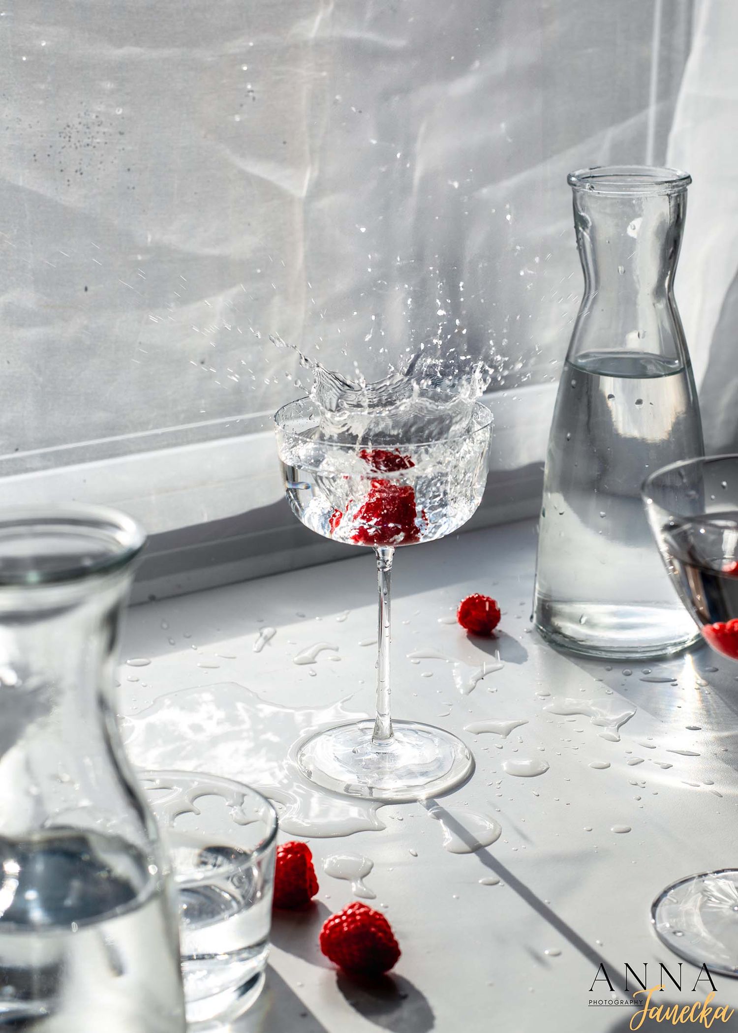 Crystal wine glass creating a splash with red berries and water on a bright windowsill.