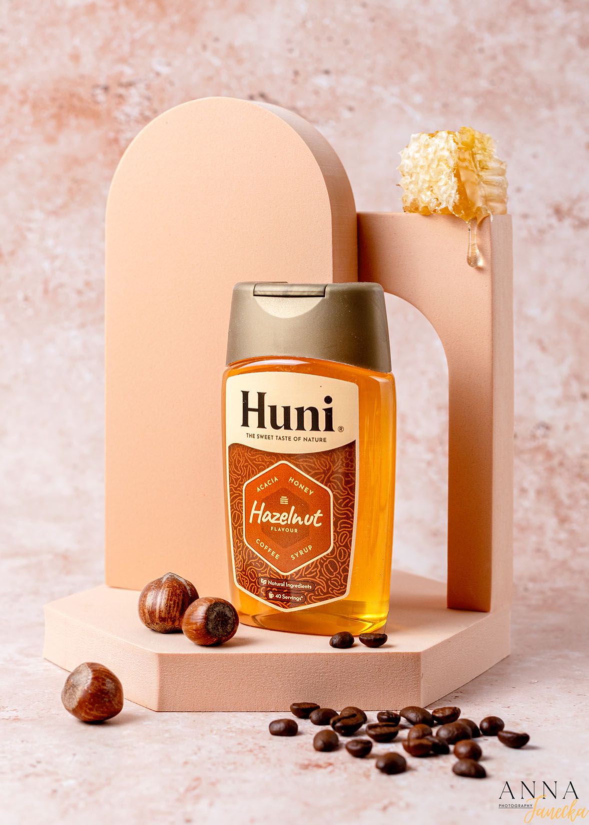 Bottle of Huni hazelnut syrup with coffee beans and hazelnuts on pink display stand.