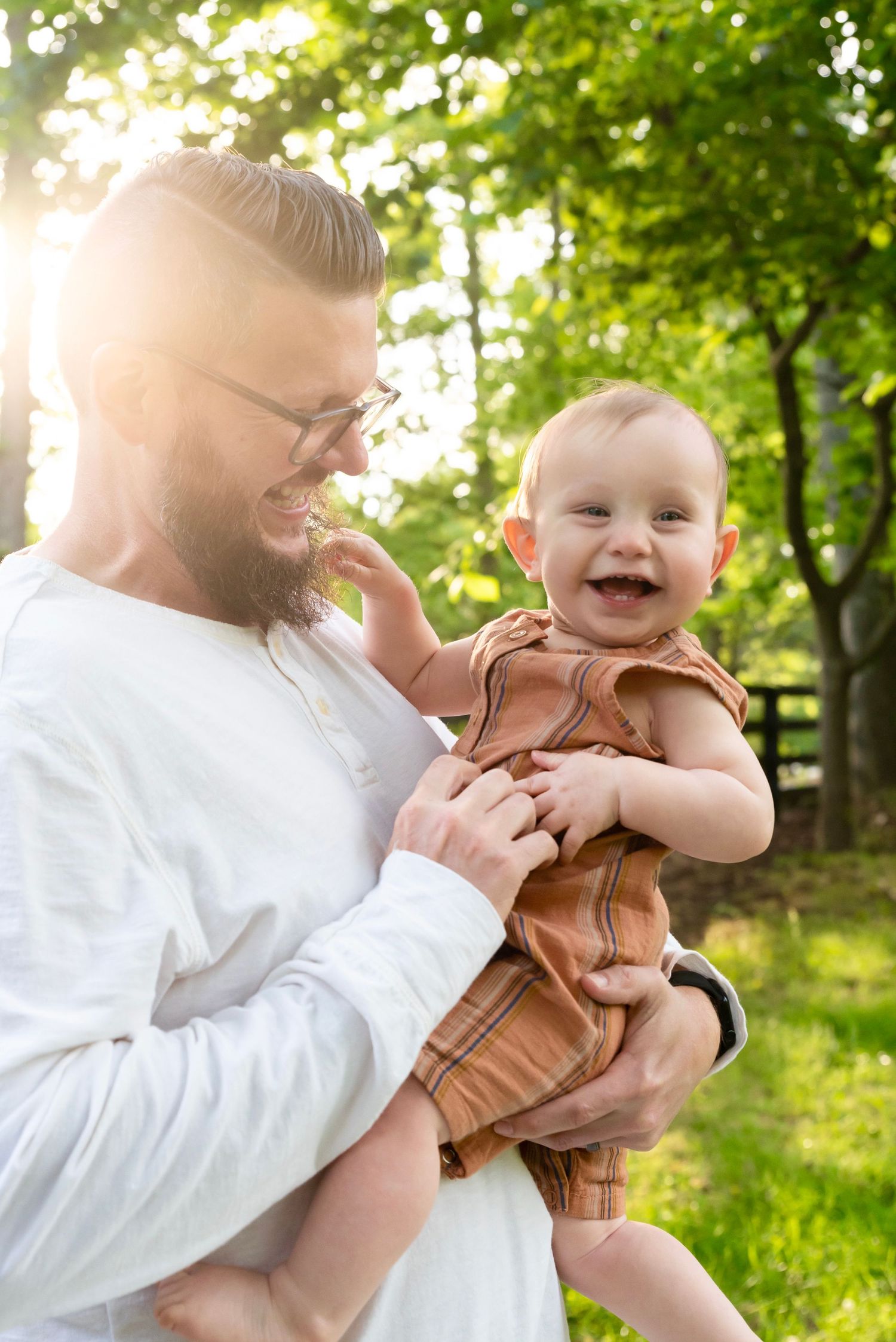 A parent in a white shirt holds and kisses a laughing baby in a brown outfit outdoors in a sunny garden setting.