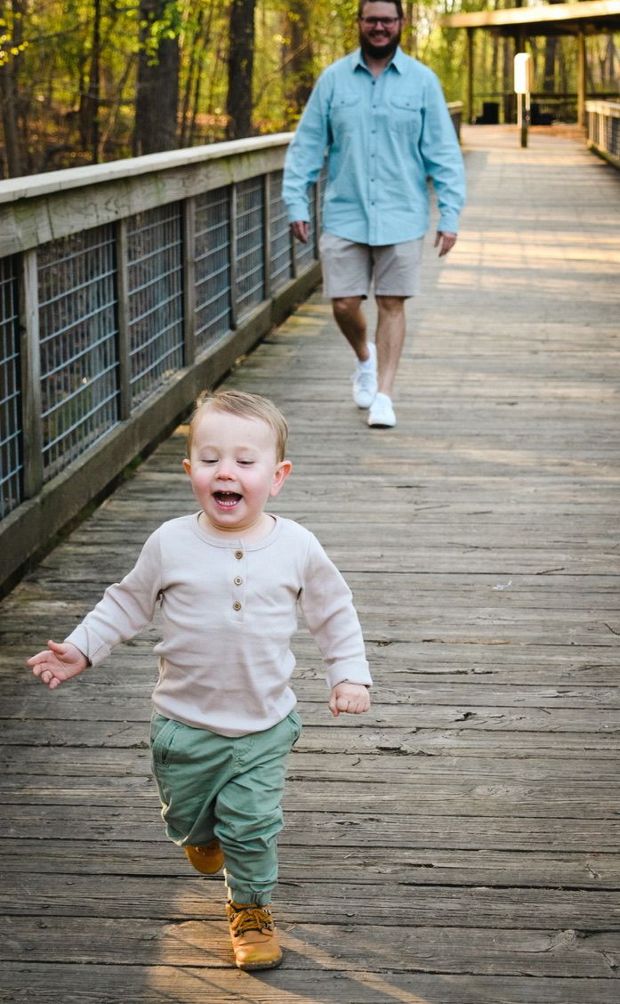 Toddler in beige shirt walking excitedly on wooden boardwalk through park.