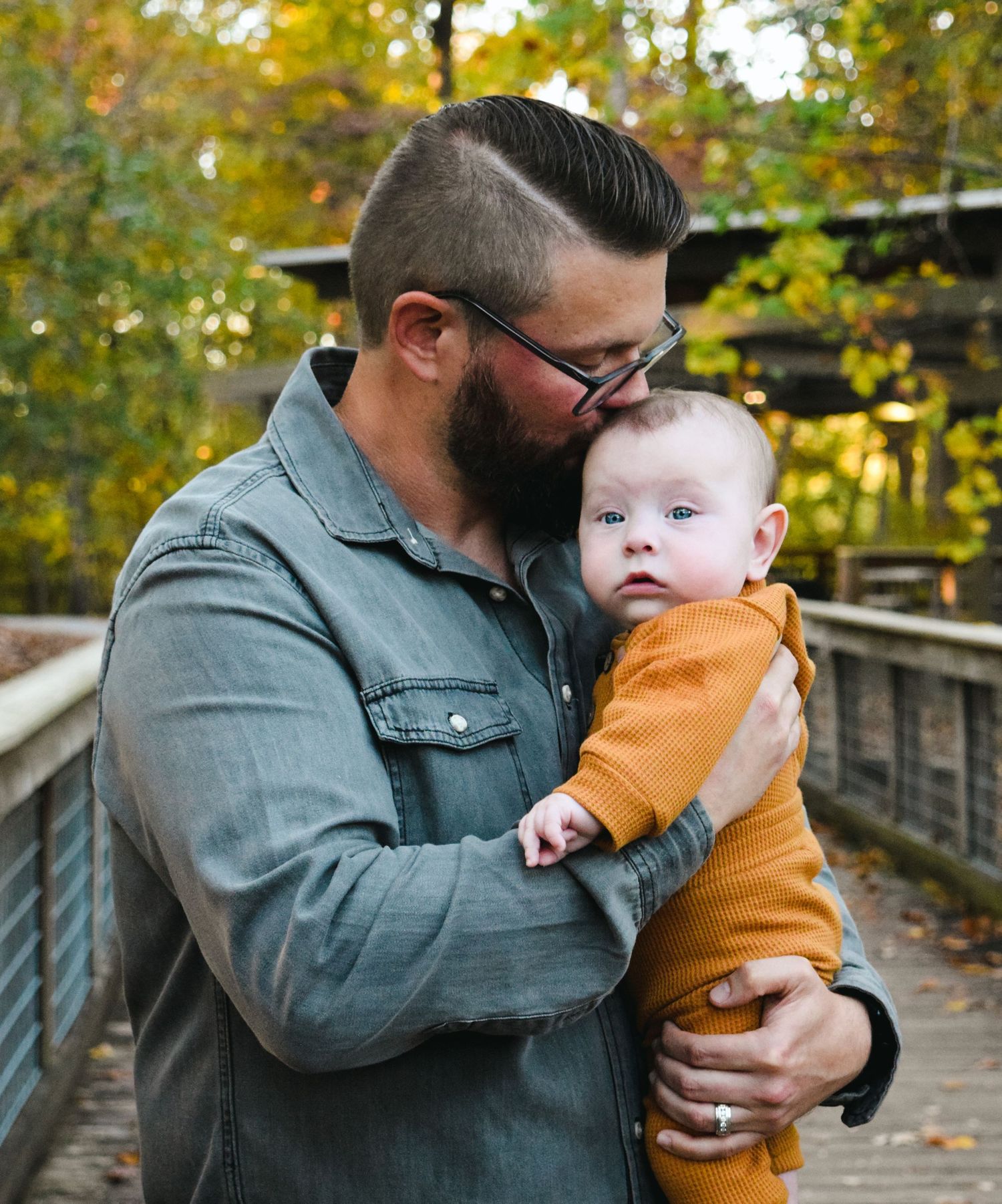 A parent wearing a denim jacket shares tender moments with a baby in an orange outfit on a wooden bridge in autumn.