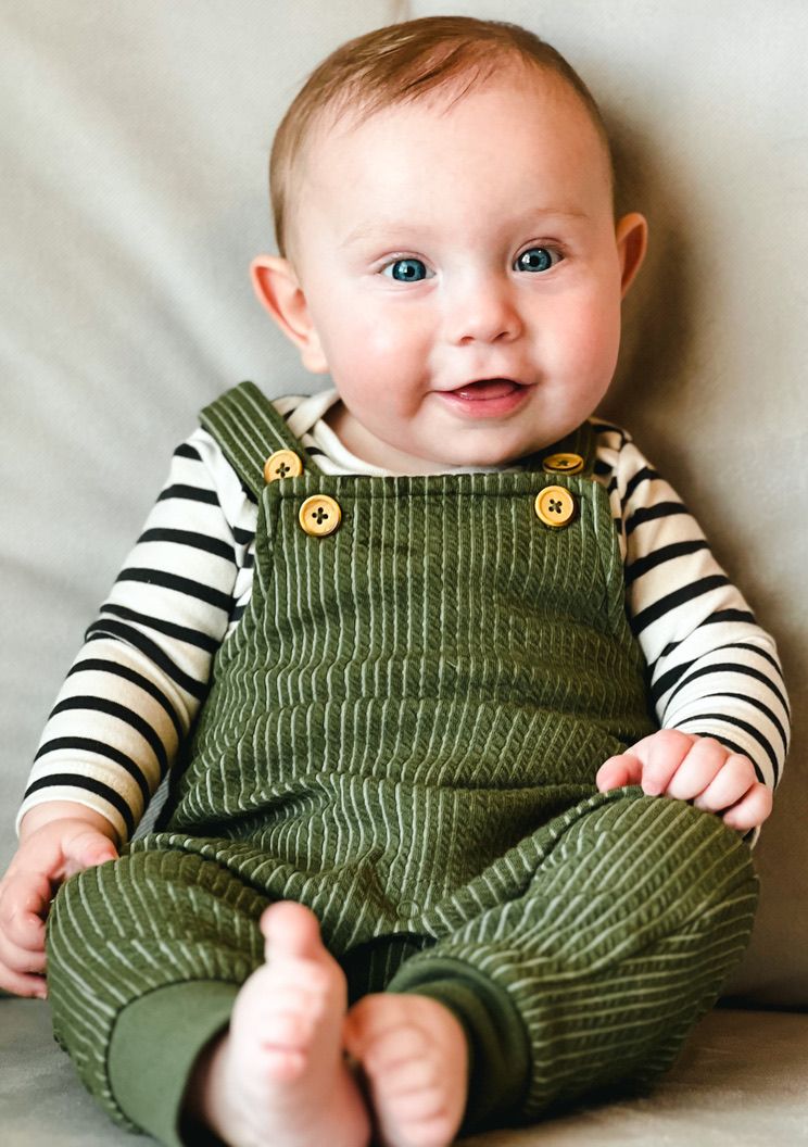 Cheerful baby wearing striped shirt and green corduroy overalls sits smiling.