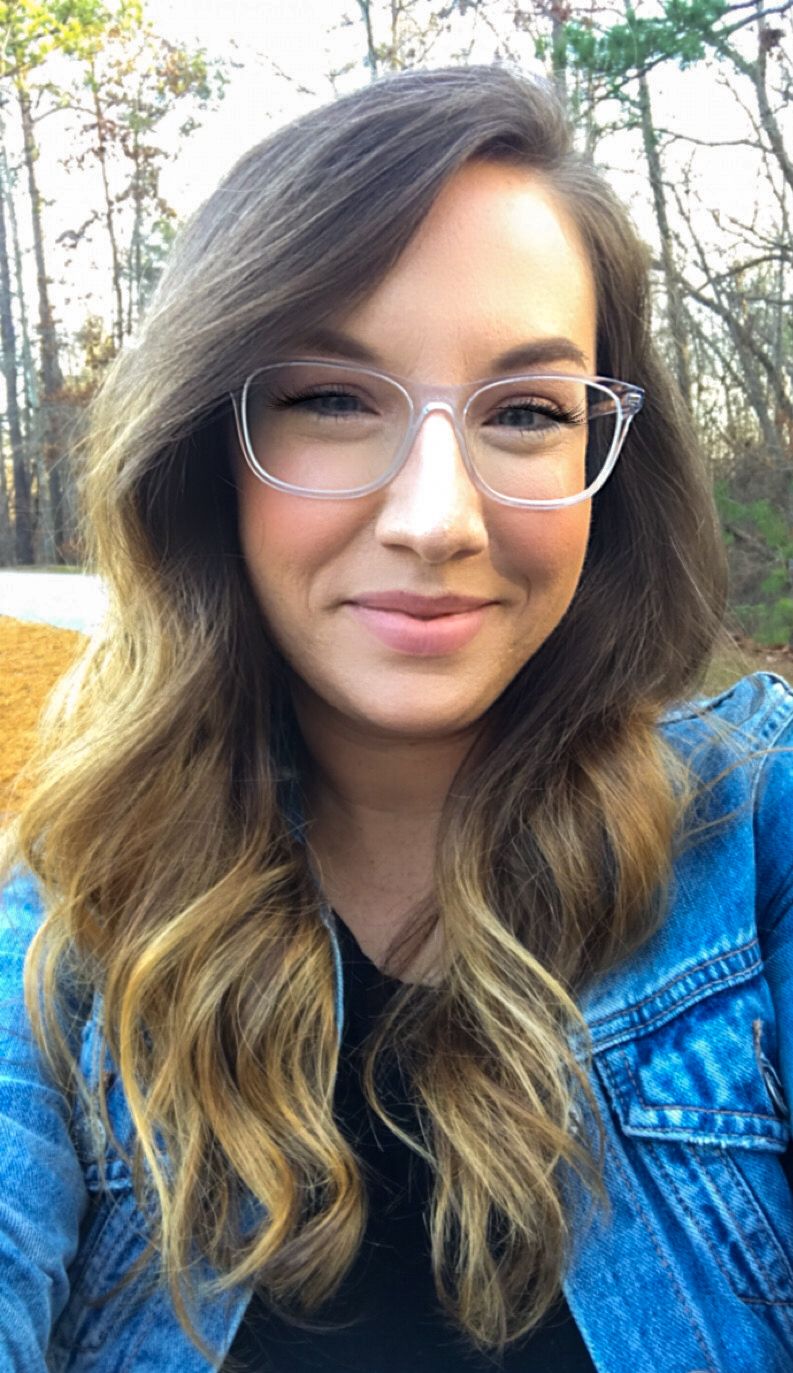 Person in denim jacket with wavy hair and glasses smiling at camera outdoors.