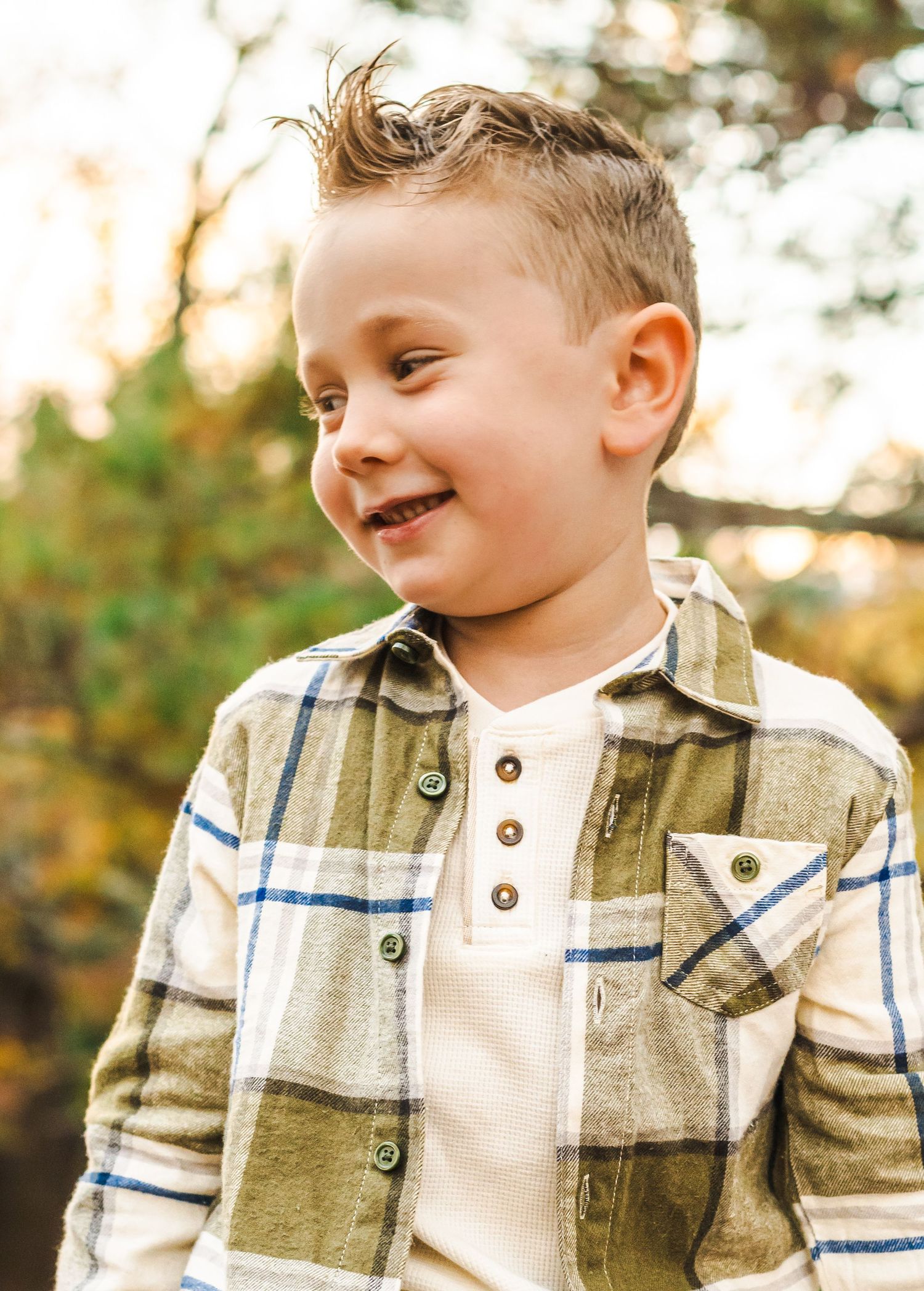 Child wearing plaid flannel button-up shirt smiling in autumn outdoor setting.