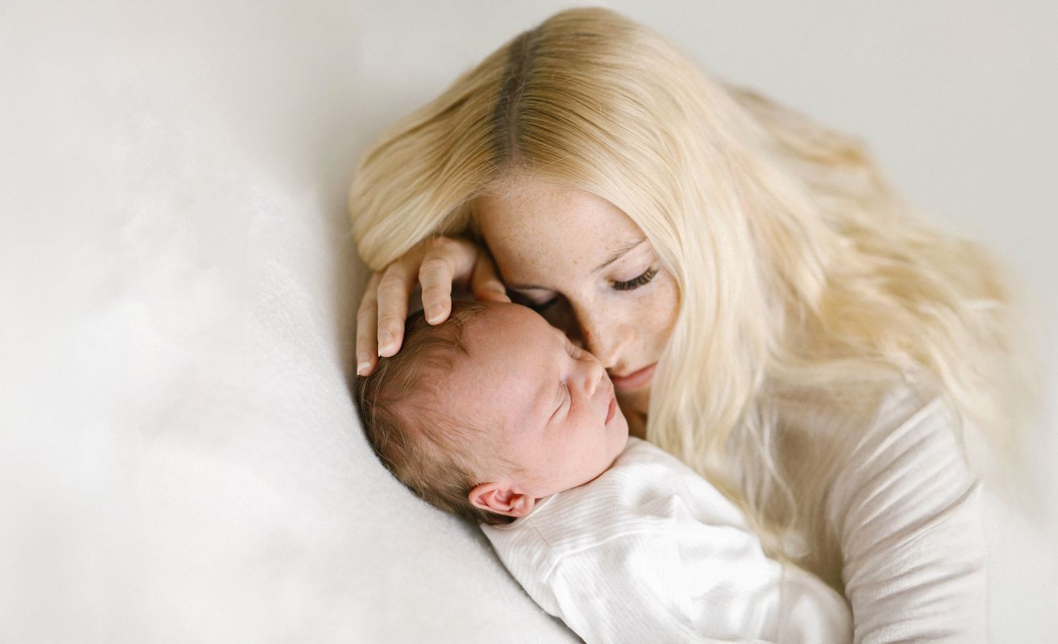 A blonde mother tenderly kisses her sleeping newborn baby wrapped in white, sharing a peaceful bonding moment.
