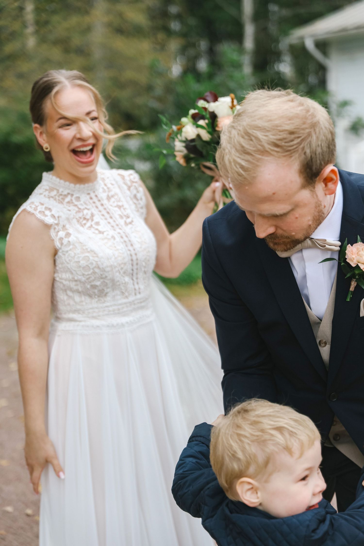 Joyful bride in lace wedding dress laughing with groom in navy suit and young child outdoors.