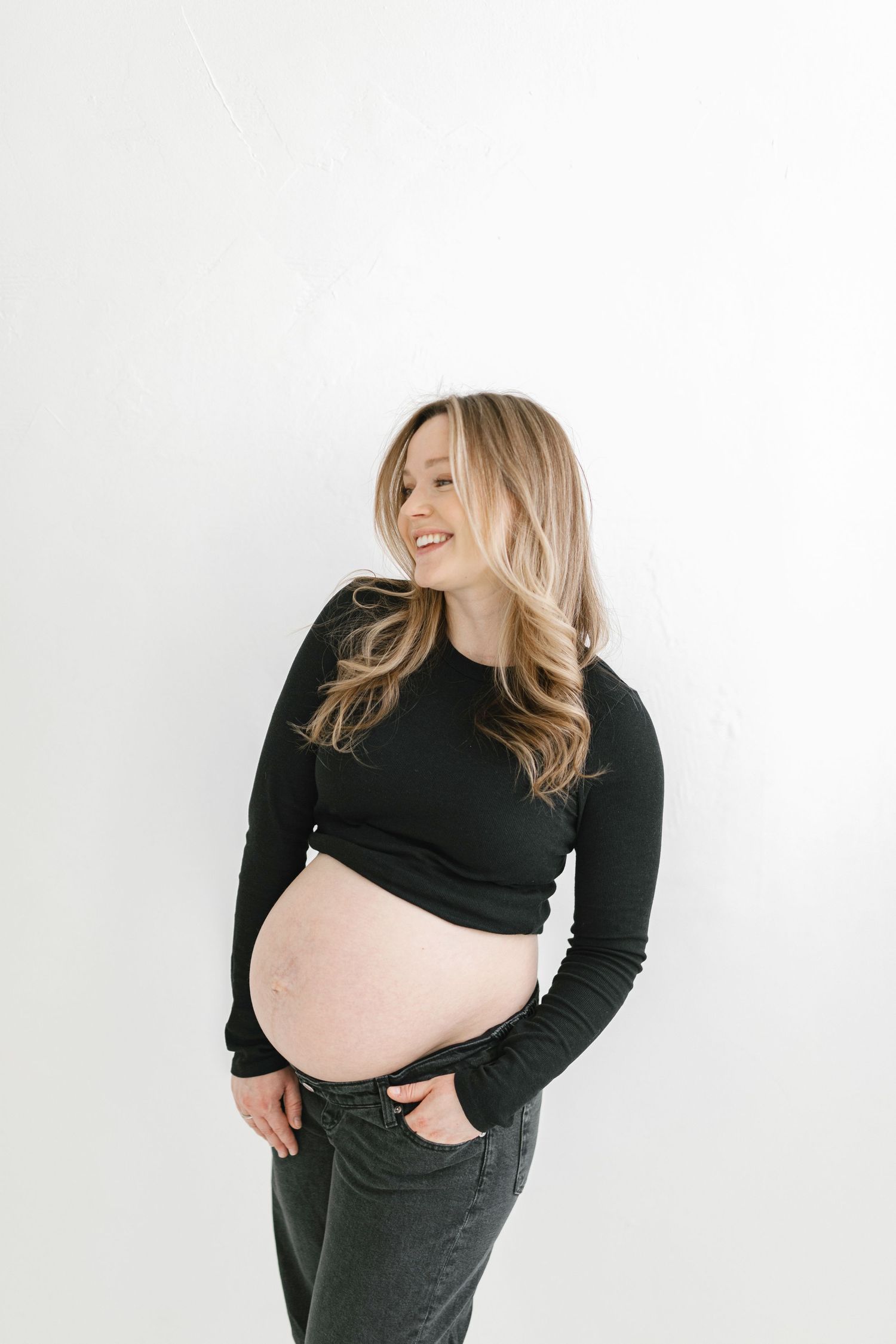 A pregnant woman in black long sleeves and dark pants poses against a white background in four sequential photos.