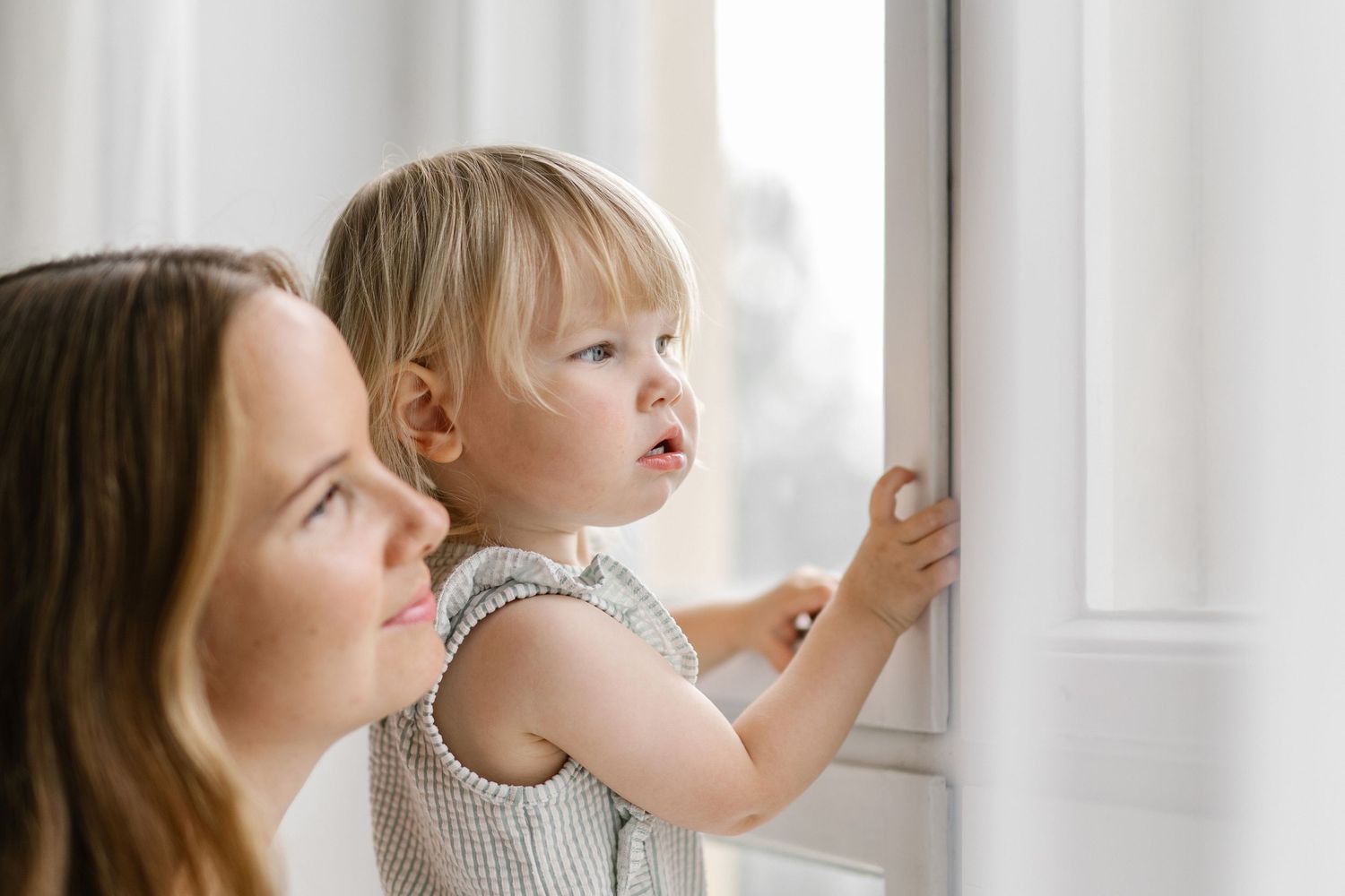 A mother holds her blonde toddler as they both look curiously out a bright window together.