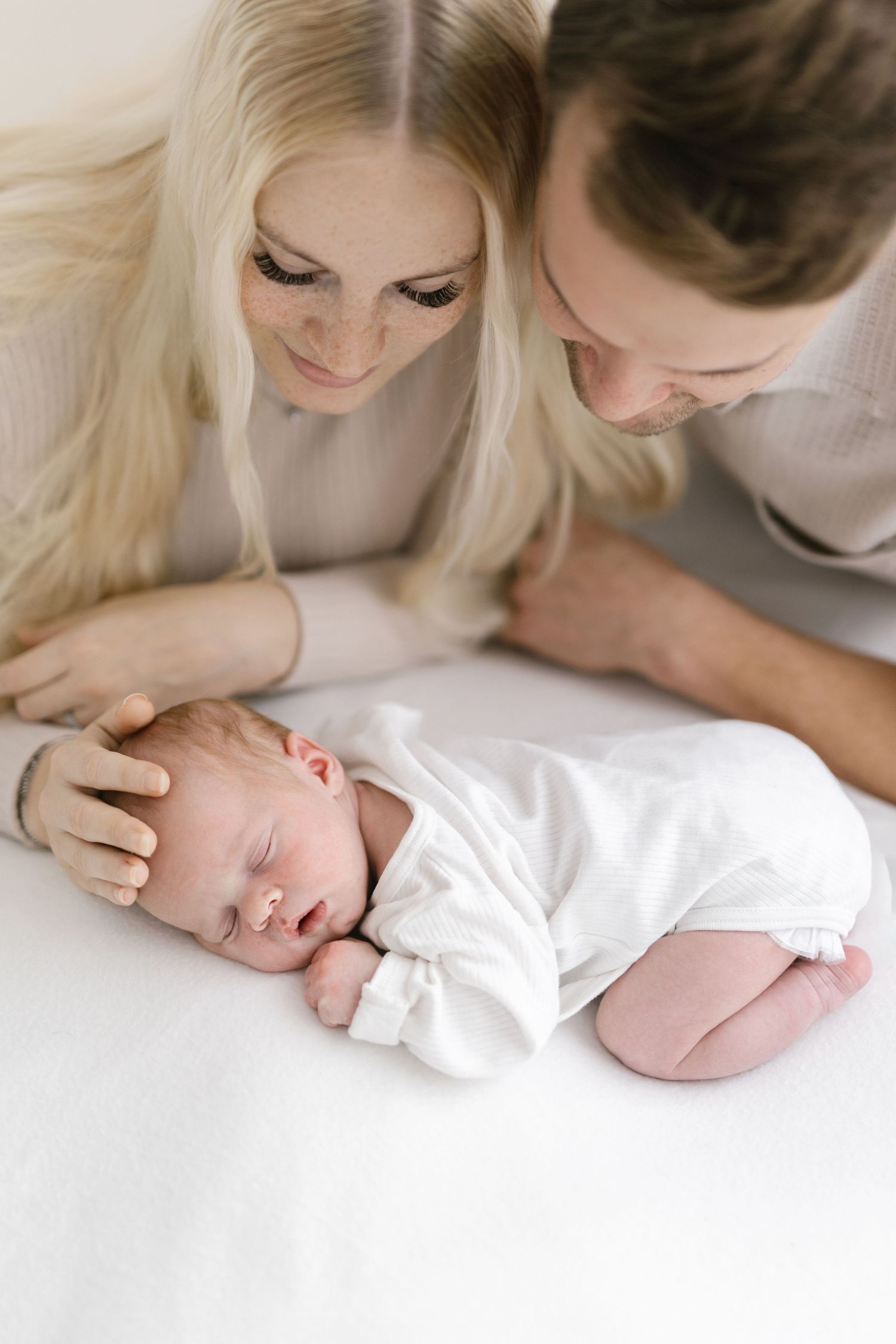 Parents lovingly watching their sleeping newborn baby on white bedding.
