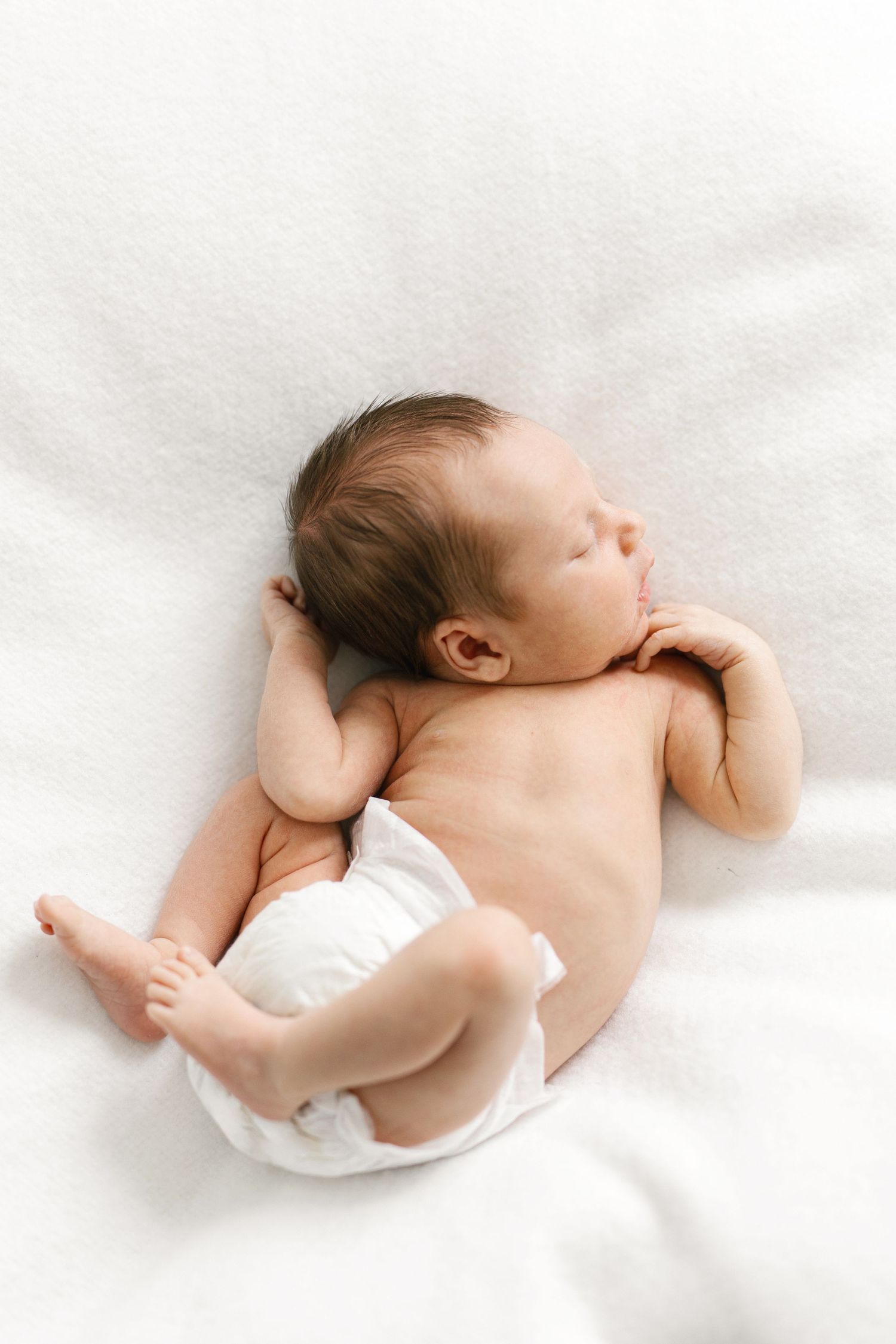 Newborn baby sleeping peacefully on white blanket, wearing diaper, with dark hair and tiny feet visible.