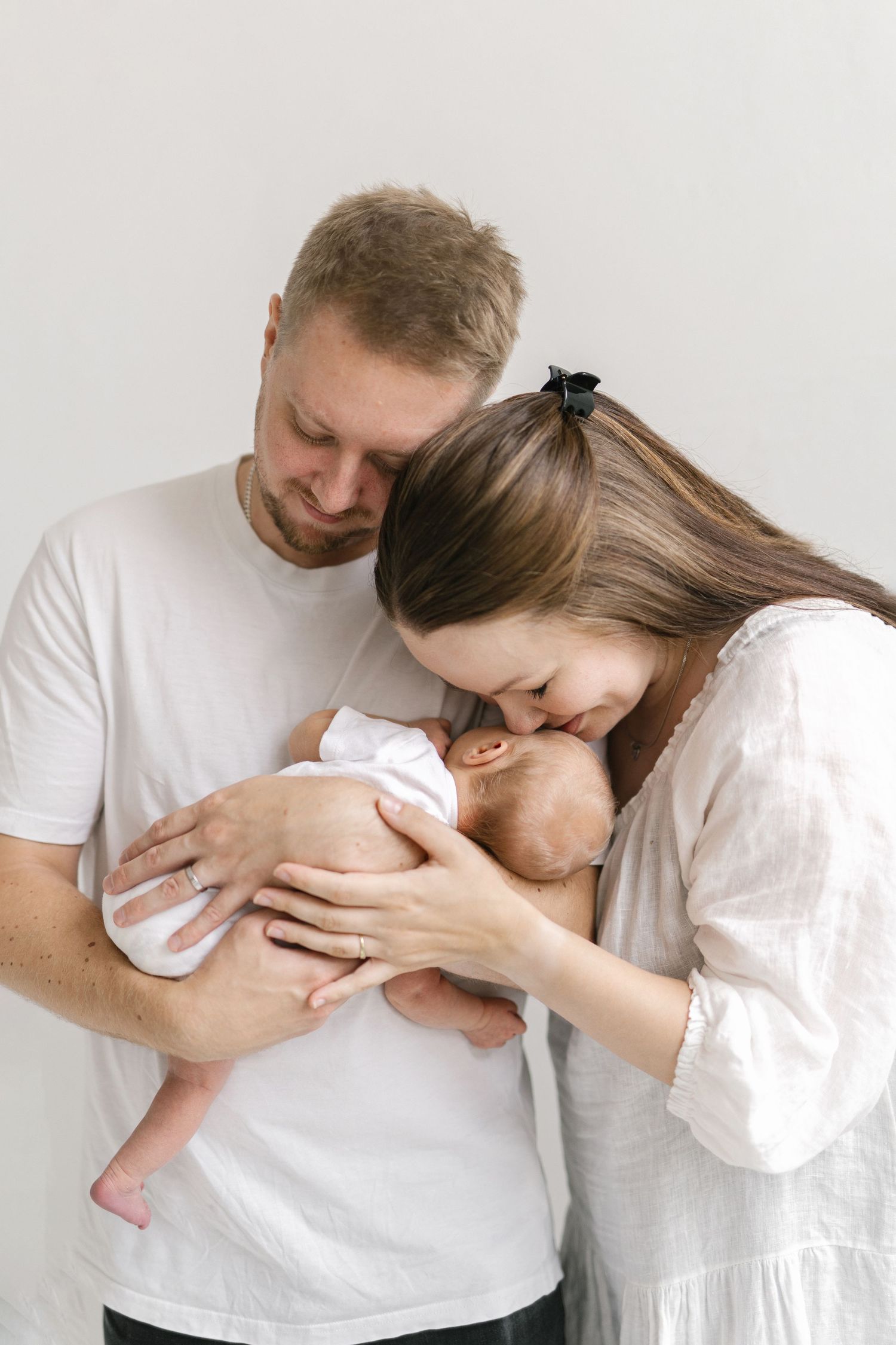 Young parents in white clothing tenderly kissing and holding their newborn baby against a light background.