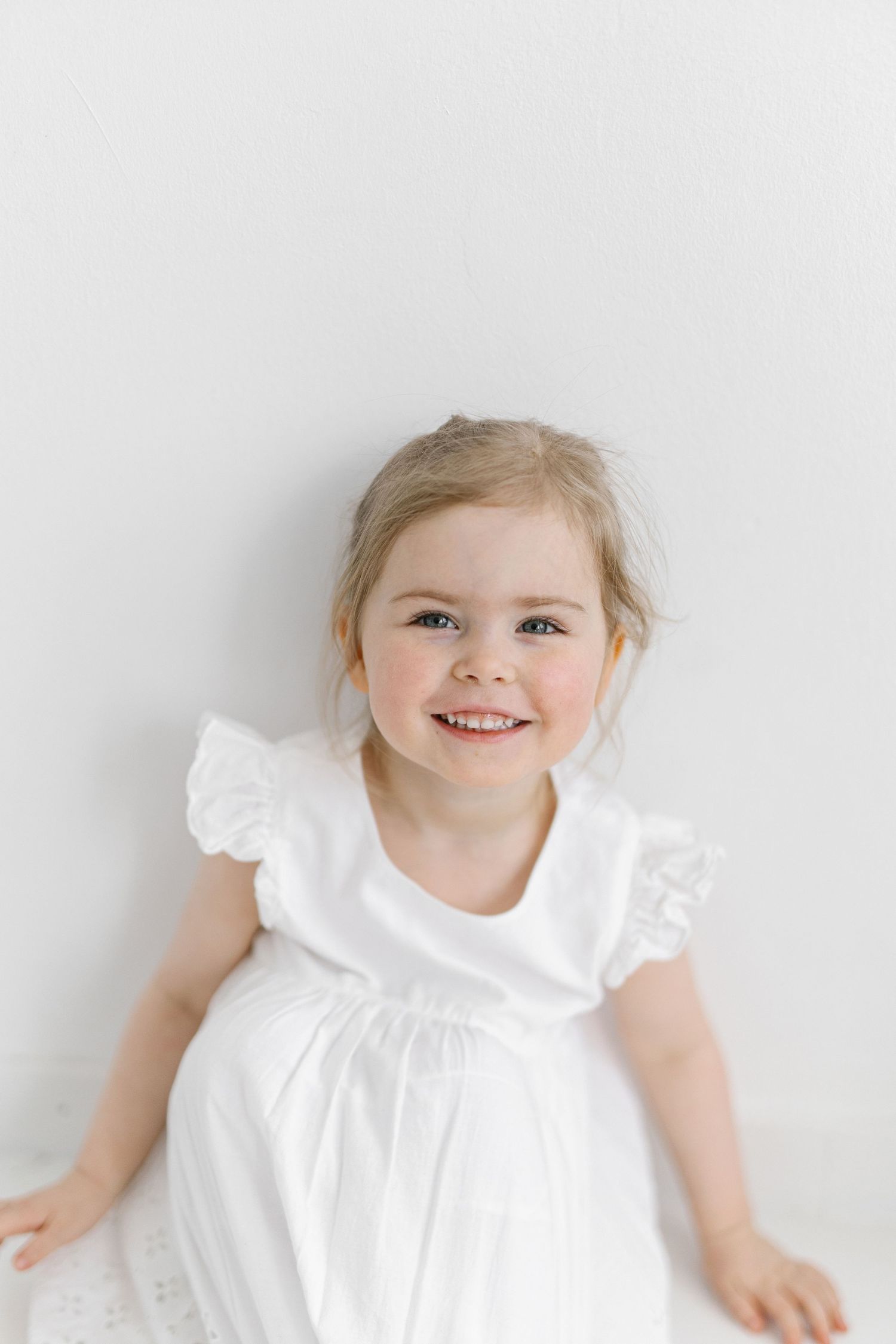 Smiling toddler girl with blonde hair wearing a white ruffle dress, sitting against a white background.