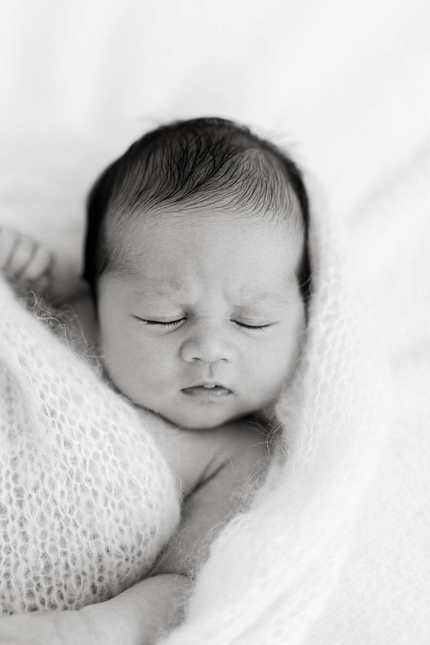 Black and white newborn portrait of a sleeping baby with dark hair wrapped in a soft knit blanket.