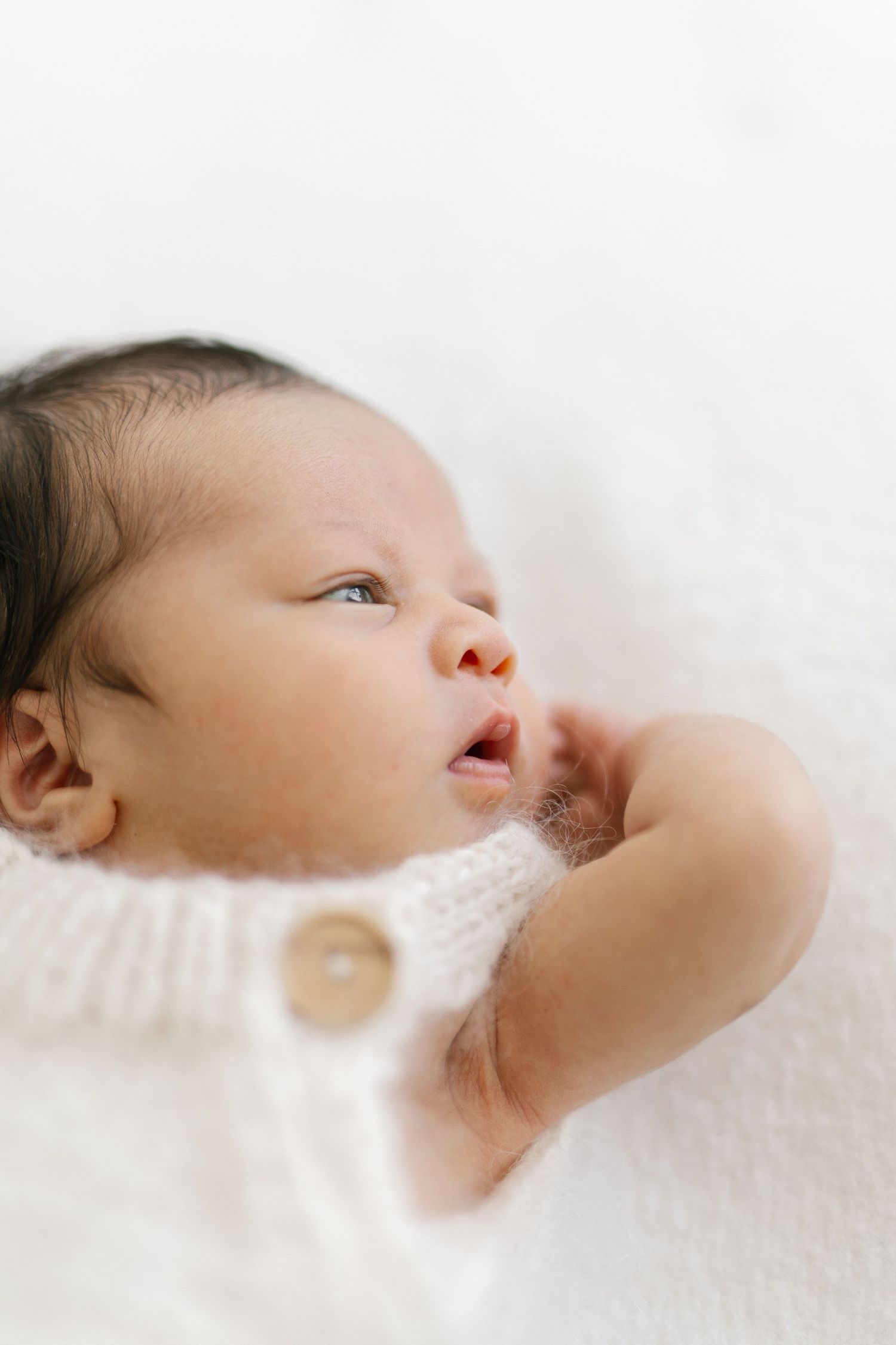 A newborn baby in a white knit romper lies on a soft white blanket, gazing upward with tiny fingers near their mouth.