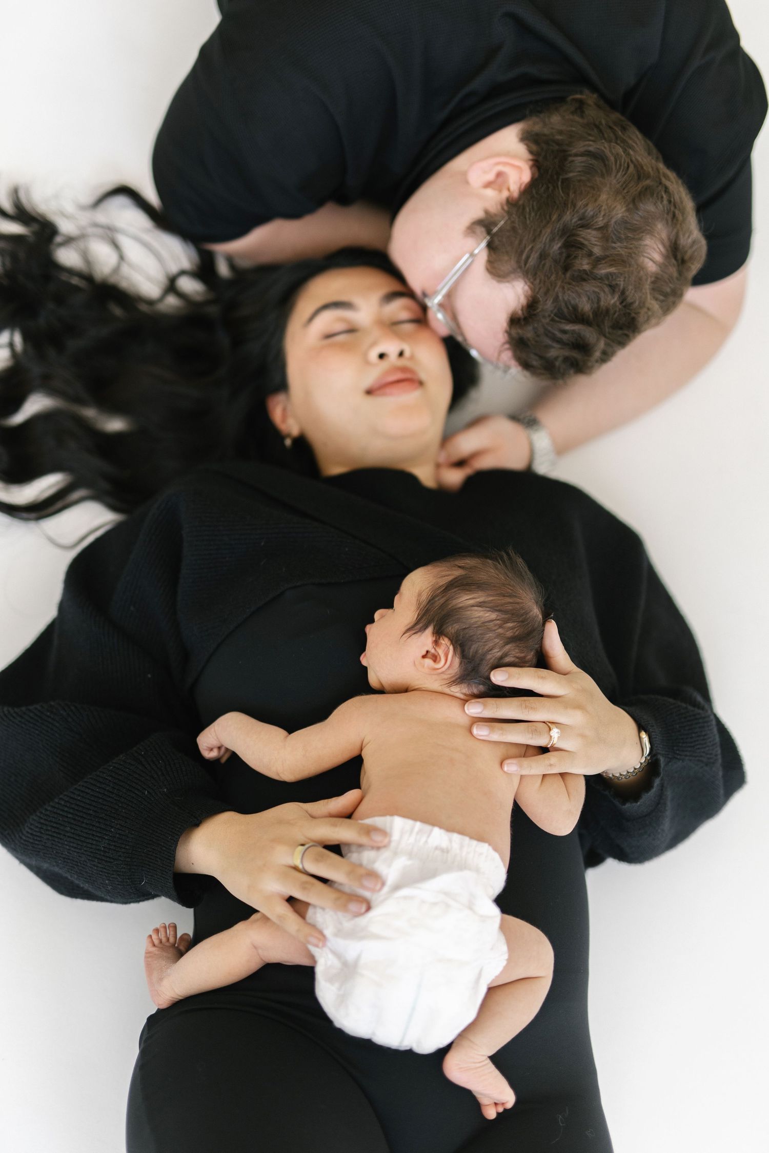 New parents in black outfits lying down with their newborn baby in a diaper on a white background.