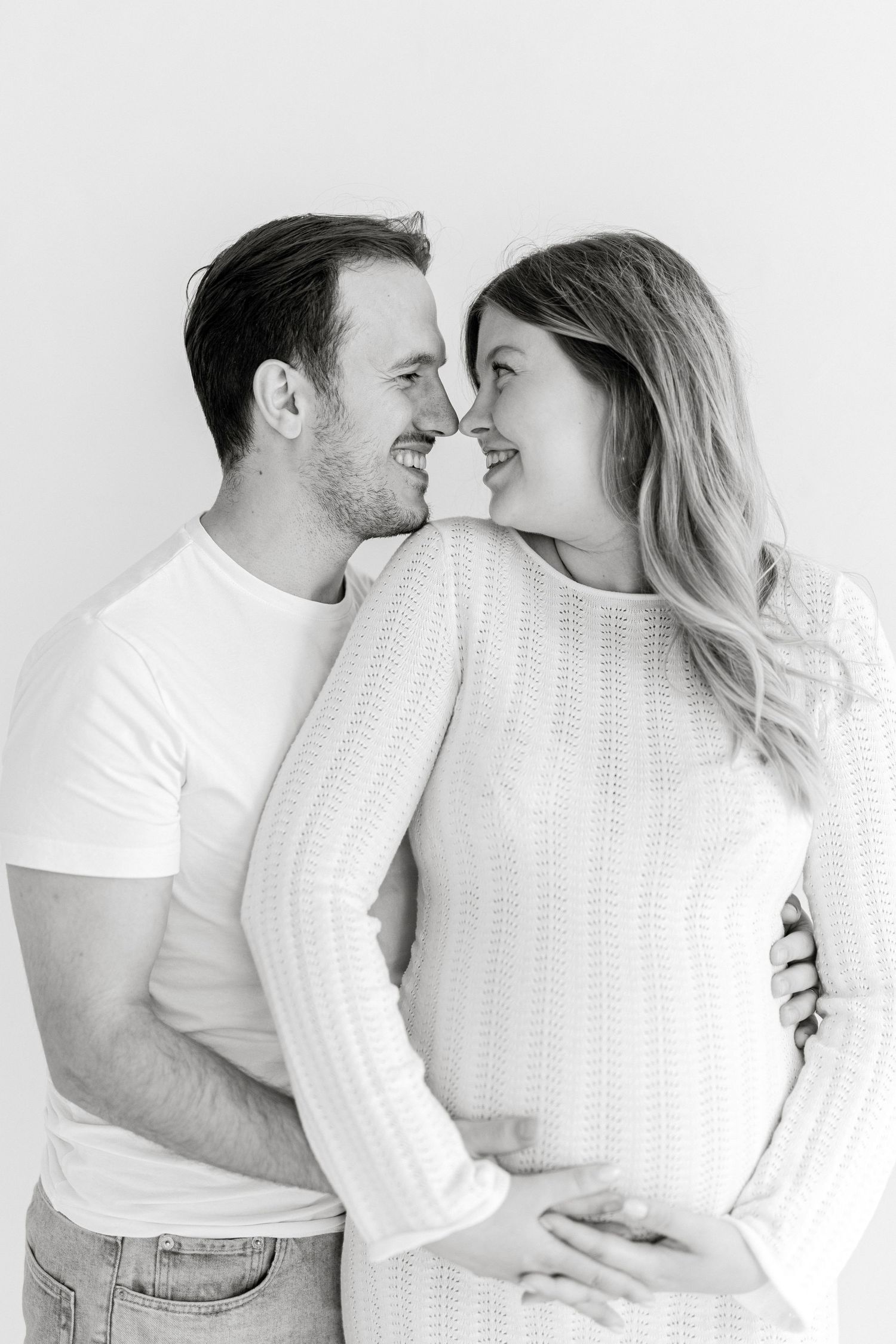 Black and white photo of a smiling couple embracing, with the woman visibly pregnant, touching noses tenderly.