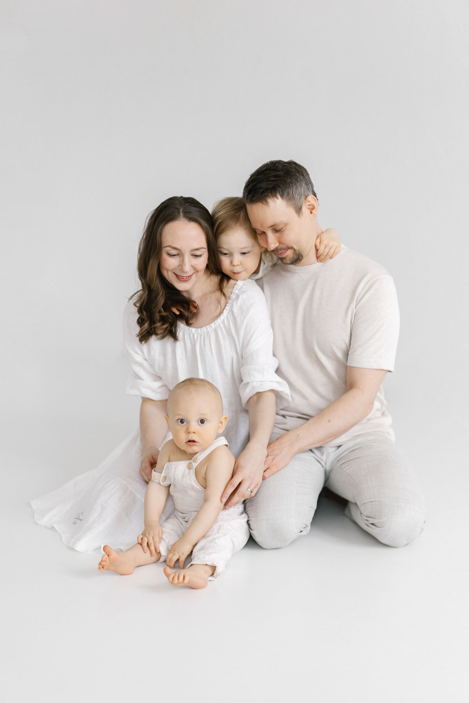 Happy family of four in white outfits posing together on white background, parents with toddler and baby.
