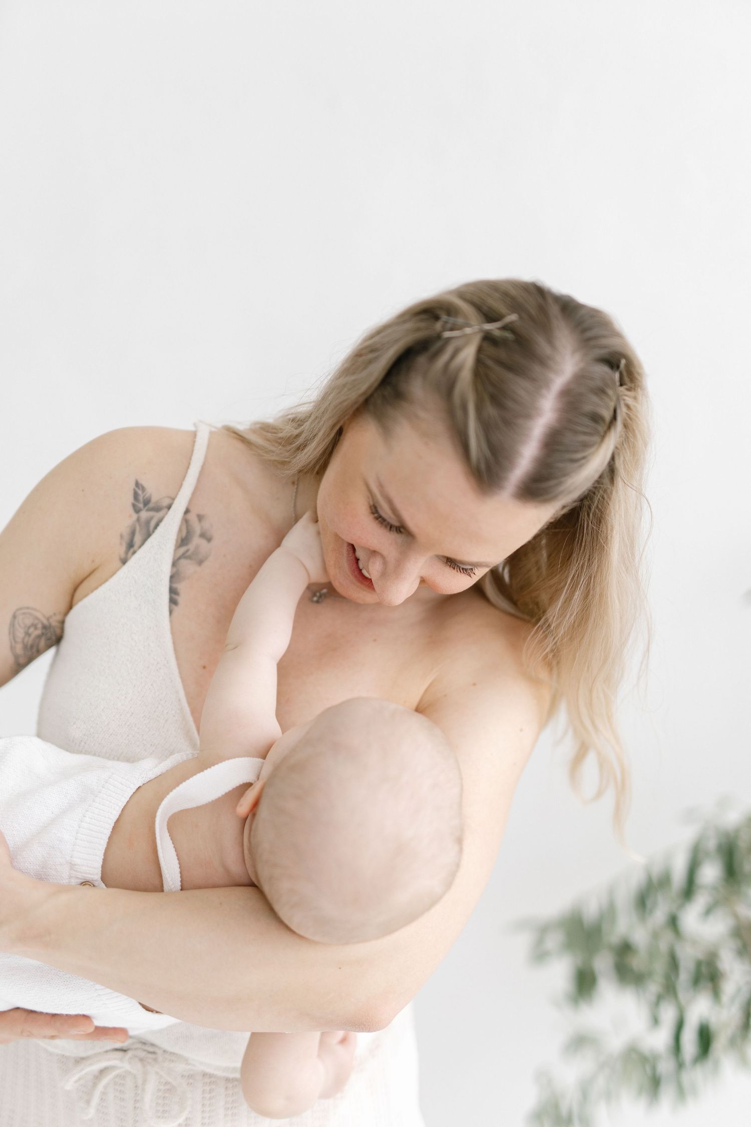 Tattooed mother breastfeeding her newborn baby, wearing a nursing bra in a bright white minimalist room.