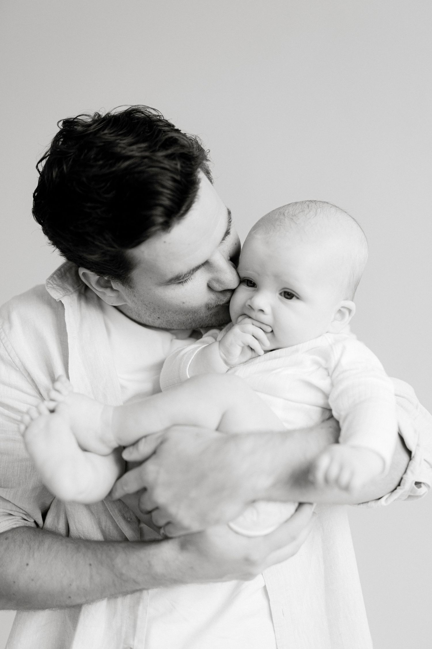 Black and white photo of a father tenderly kissing his newborn baby while cradling the infant in his arms.