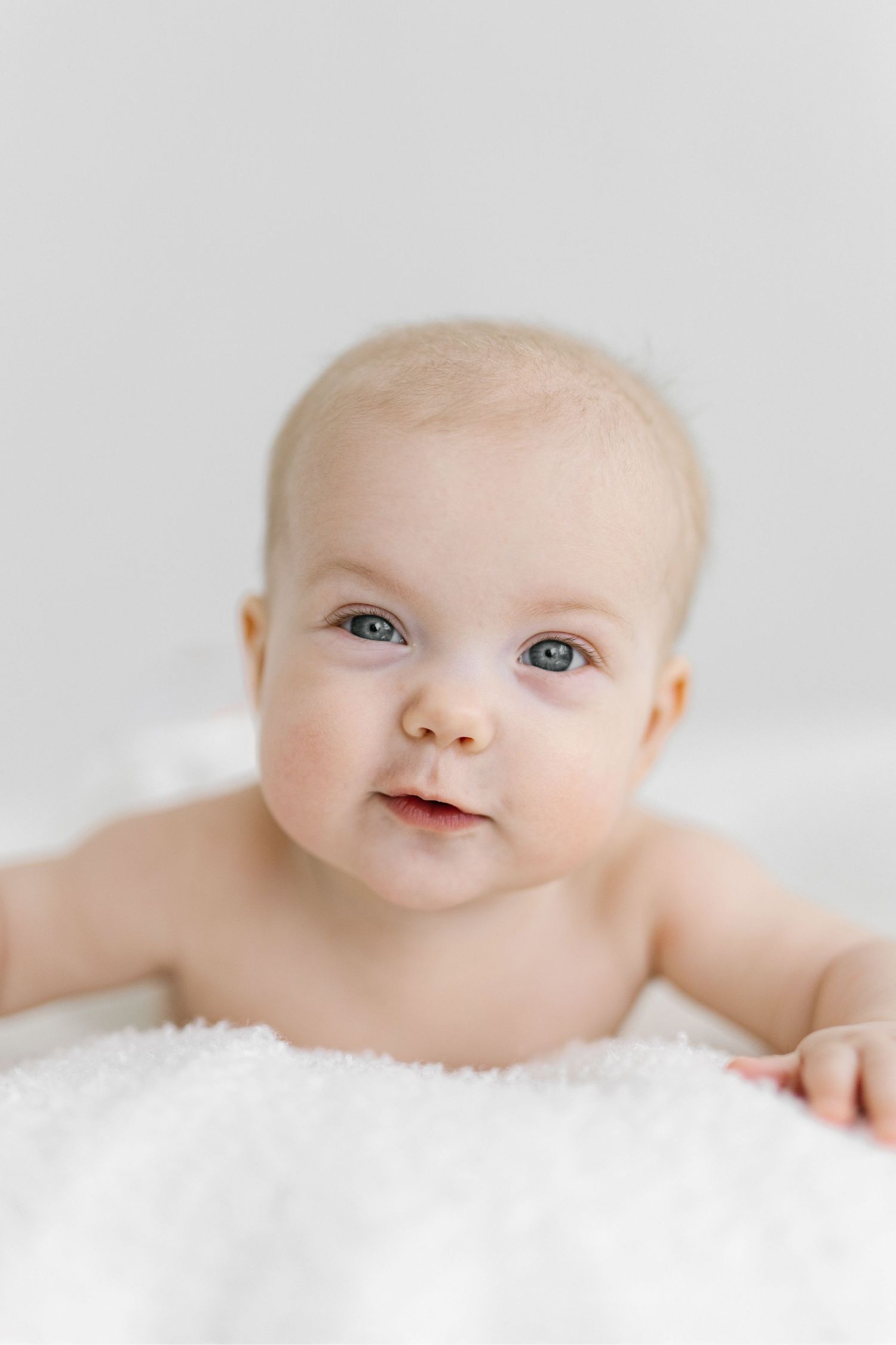 Adorable baby with blue eyes lying on a soft white fluffy blanket, looking up with a sweet curious expression.