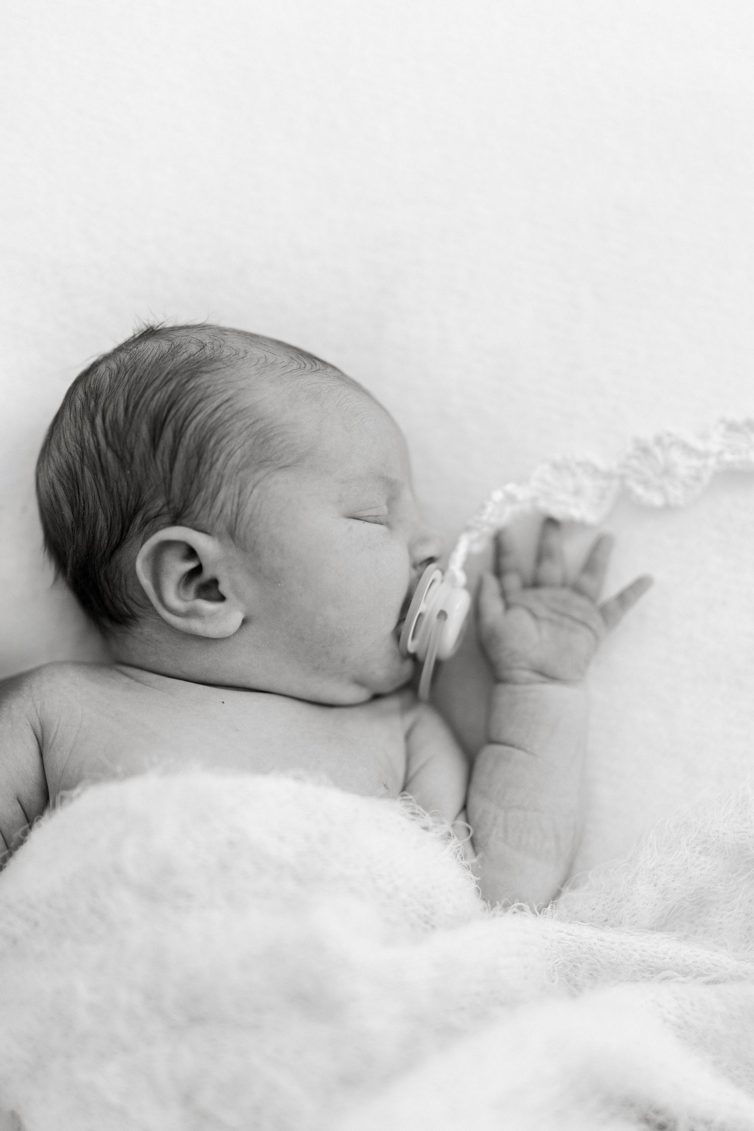 Black and white photo of a sleeping newborn baby with a pacifier, lying on a soft white blanket.