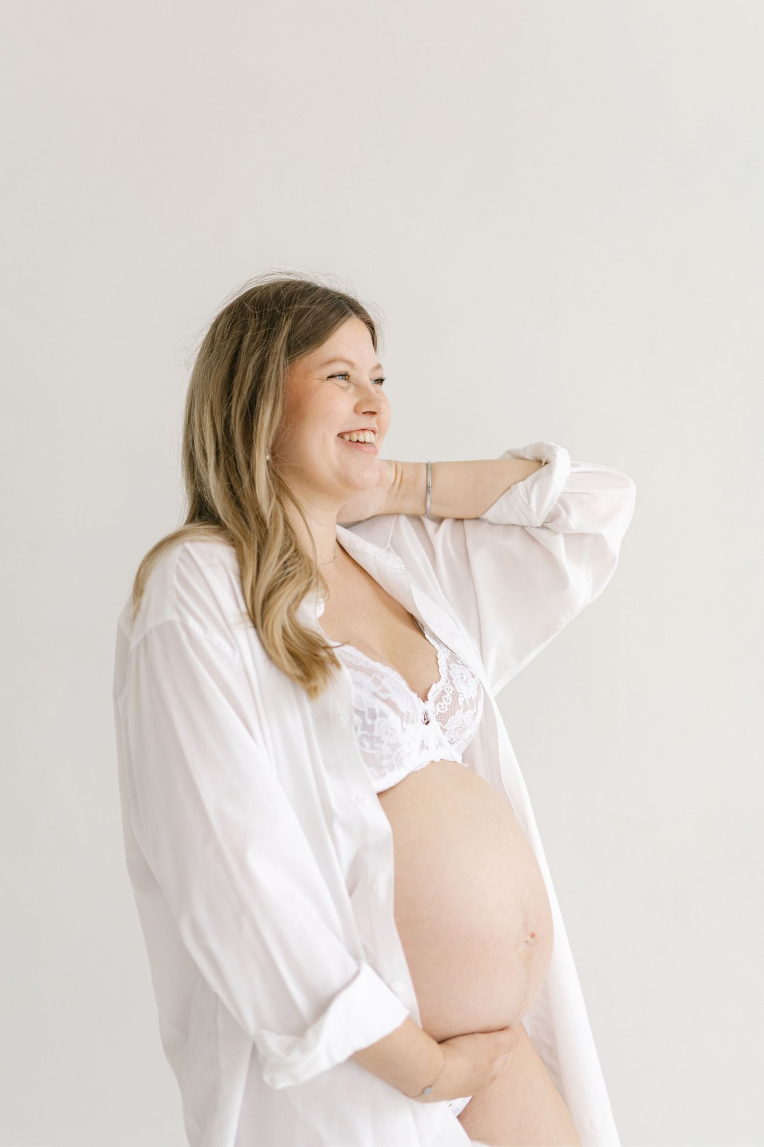 Smiling pregnant woman in white open shirt and lace bralette cradling her baby bump against a light background.